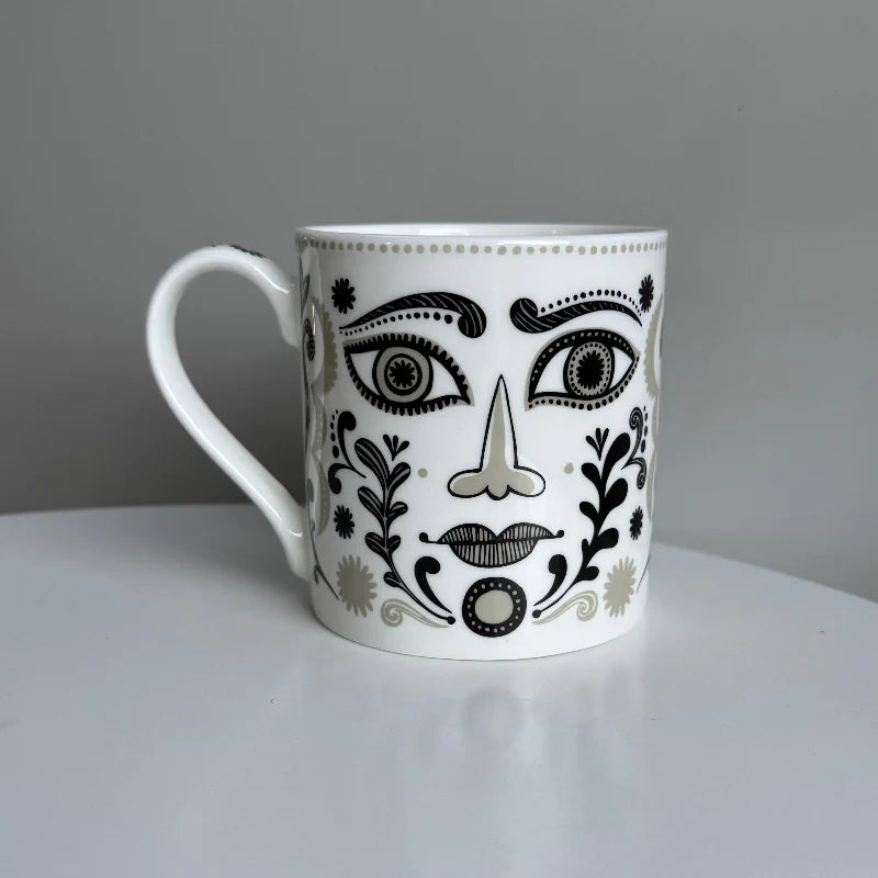 White mug with black and gray face design on a white surface with gray background