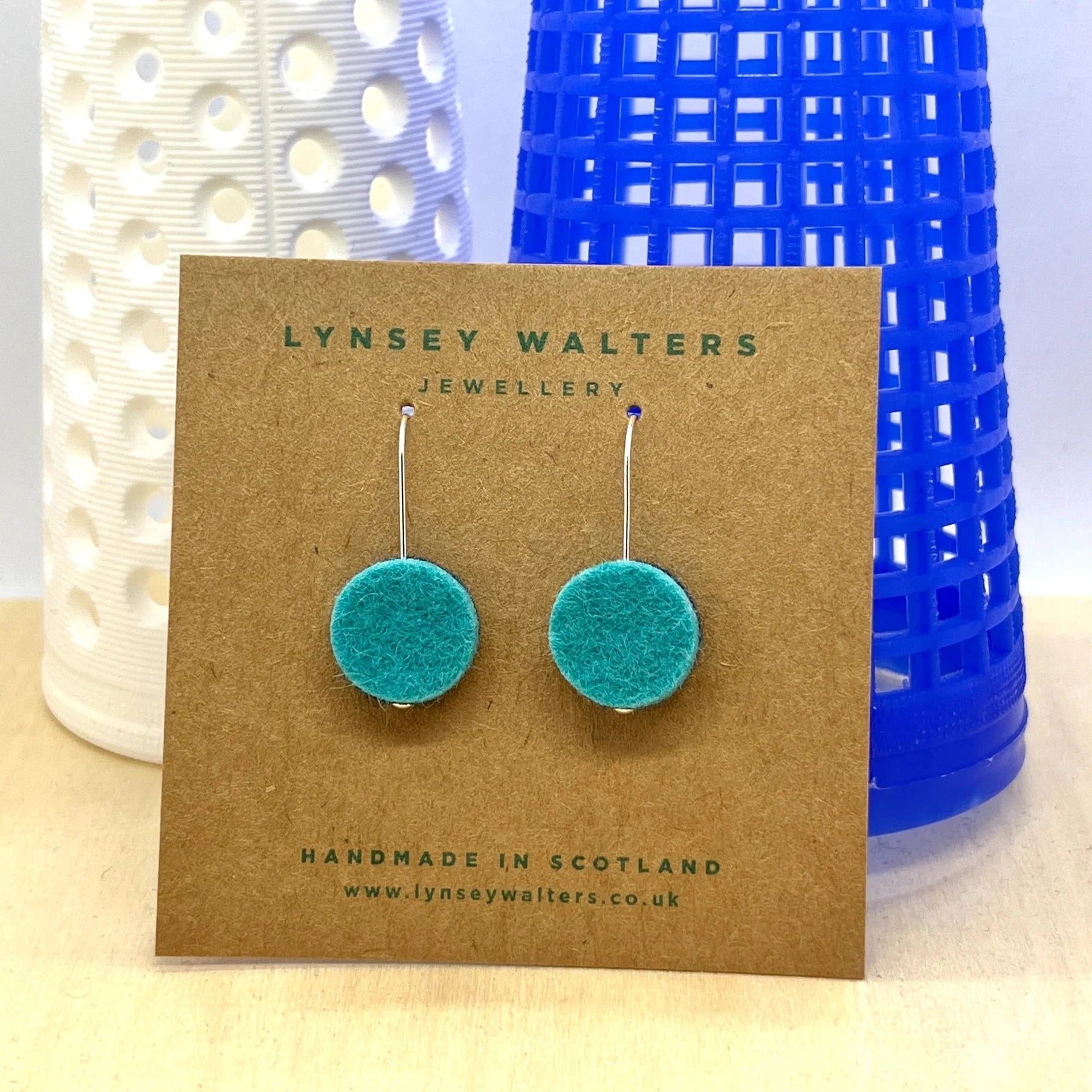 Pair of teal felt circle drop earrings with silver hooks, displayed on a brown card.