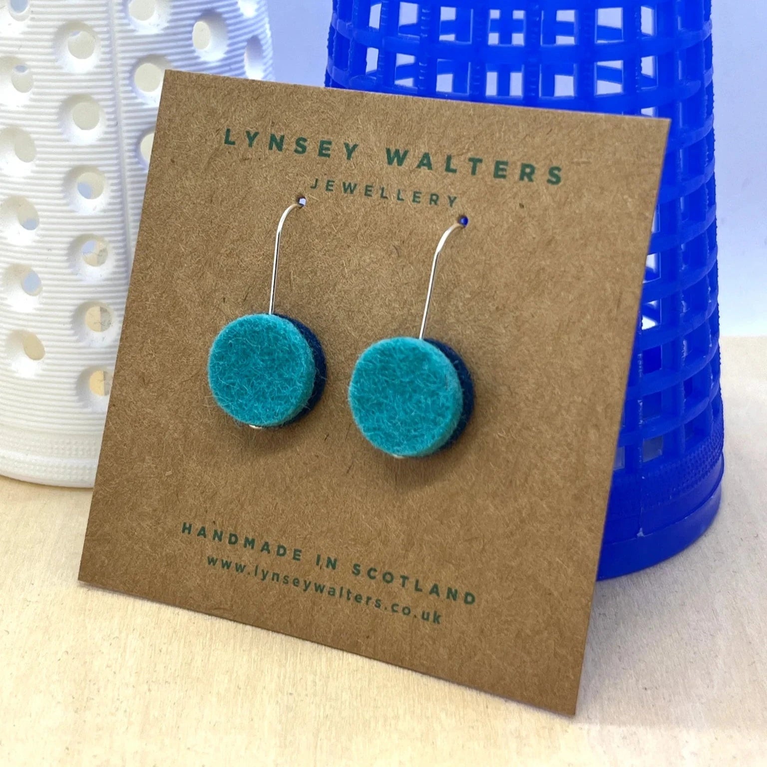 Pair of teal felt circle drop earrings with silver hooks, displayed on a brown card.