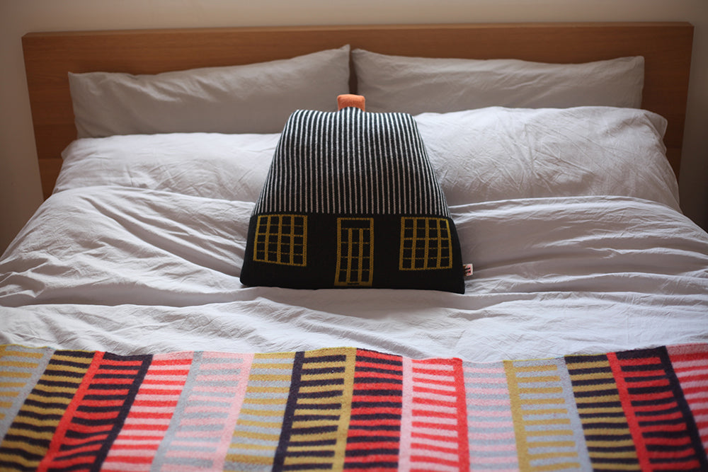 A cozy bed with white linens, featuring a black and yellow striped house pillow by Donna Wilson and a colorful patterned throw.