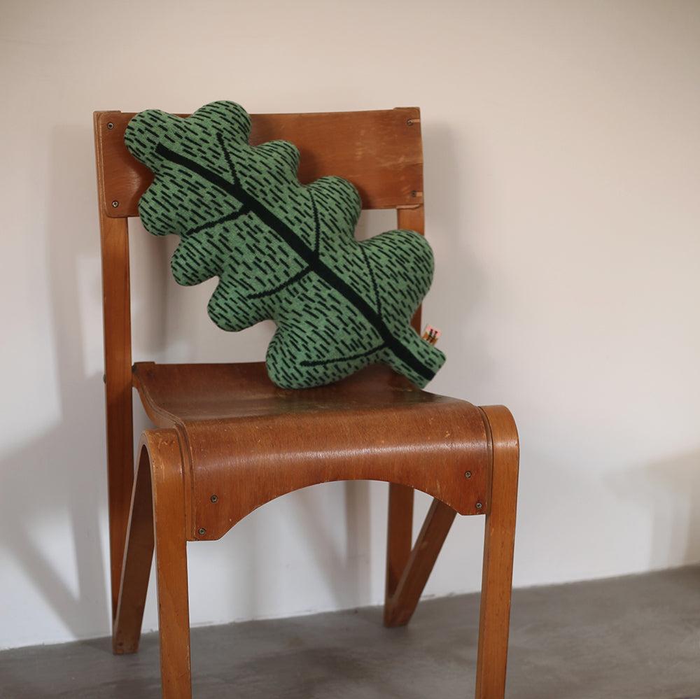 A green leaf-shaped pillow by Donna Wilson sits on a wooden chair, featuring a dark green stem and patterned texture.