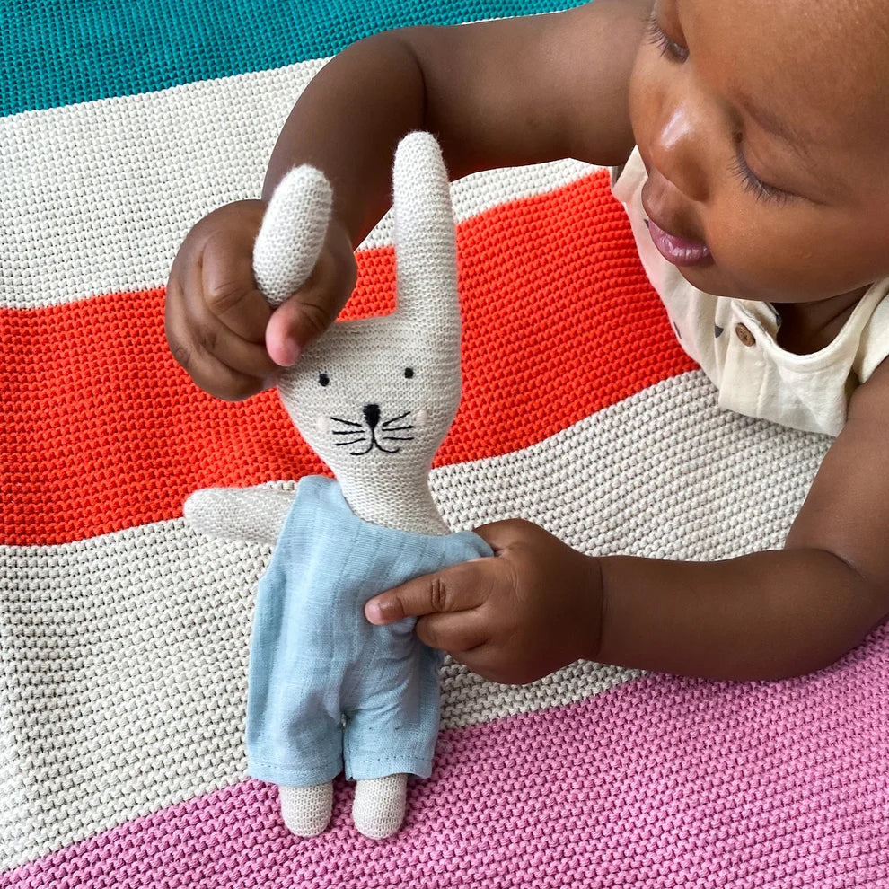 Knitted bunny toy wearing a light blue overall, held by a baby on a striped blanket.
