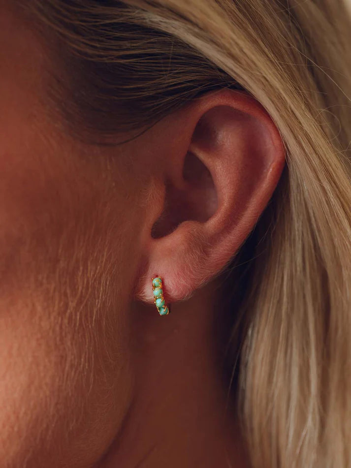 A close-up of a person's ear wearing a small curved stud earring with multiple small turquoise-blue stones set in a gold-tone band.