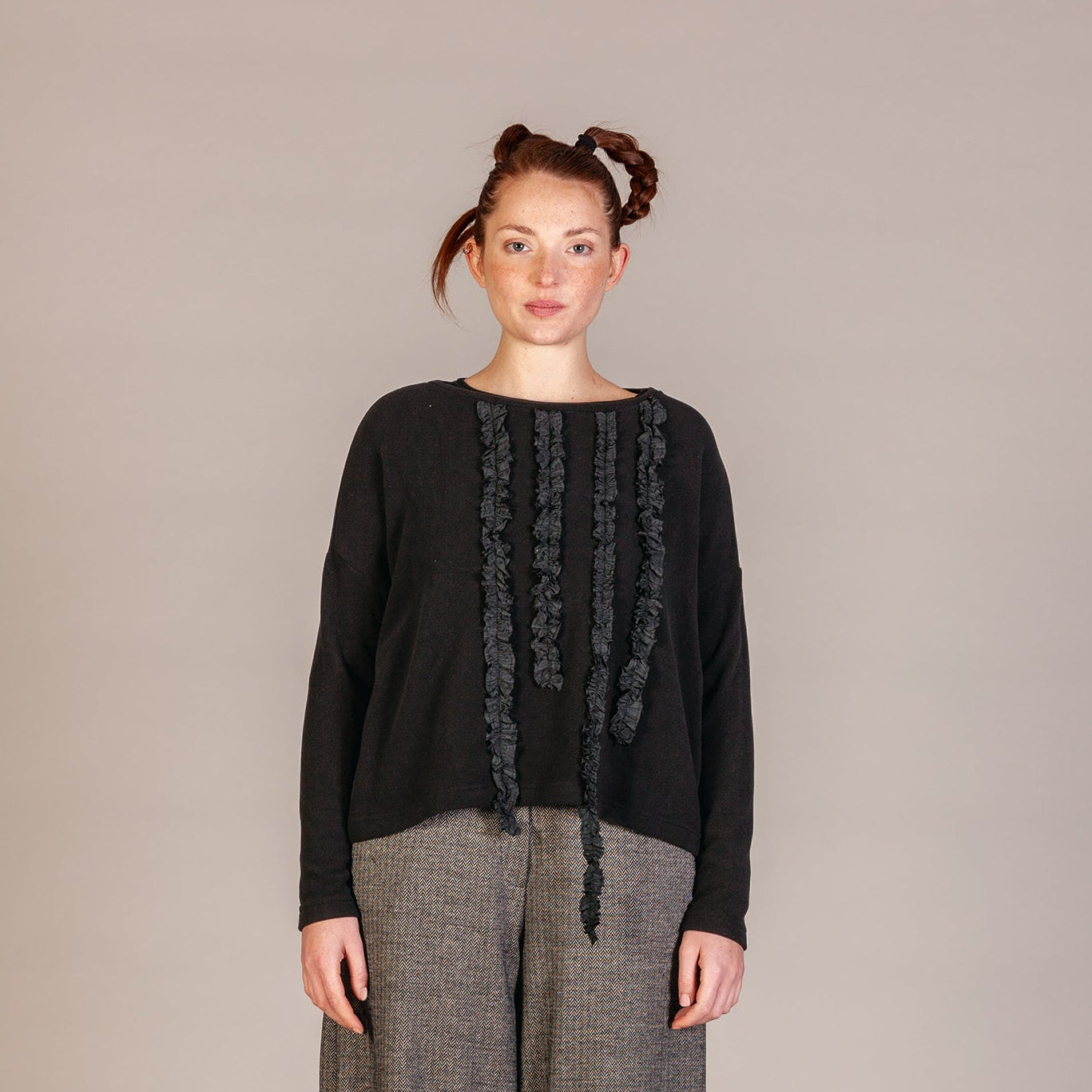 A model stands against a neutral beige studio background wearing a black long-sleeve top with vertical textured fringe details down the front, paired with loose gray wide-leg trousers and black lace-up boots.
