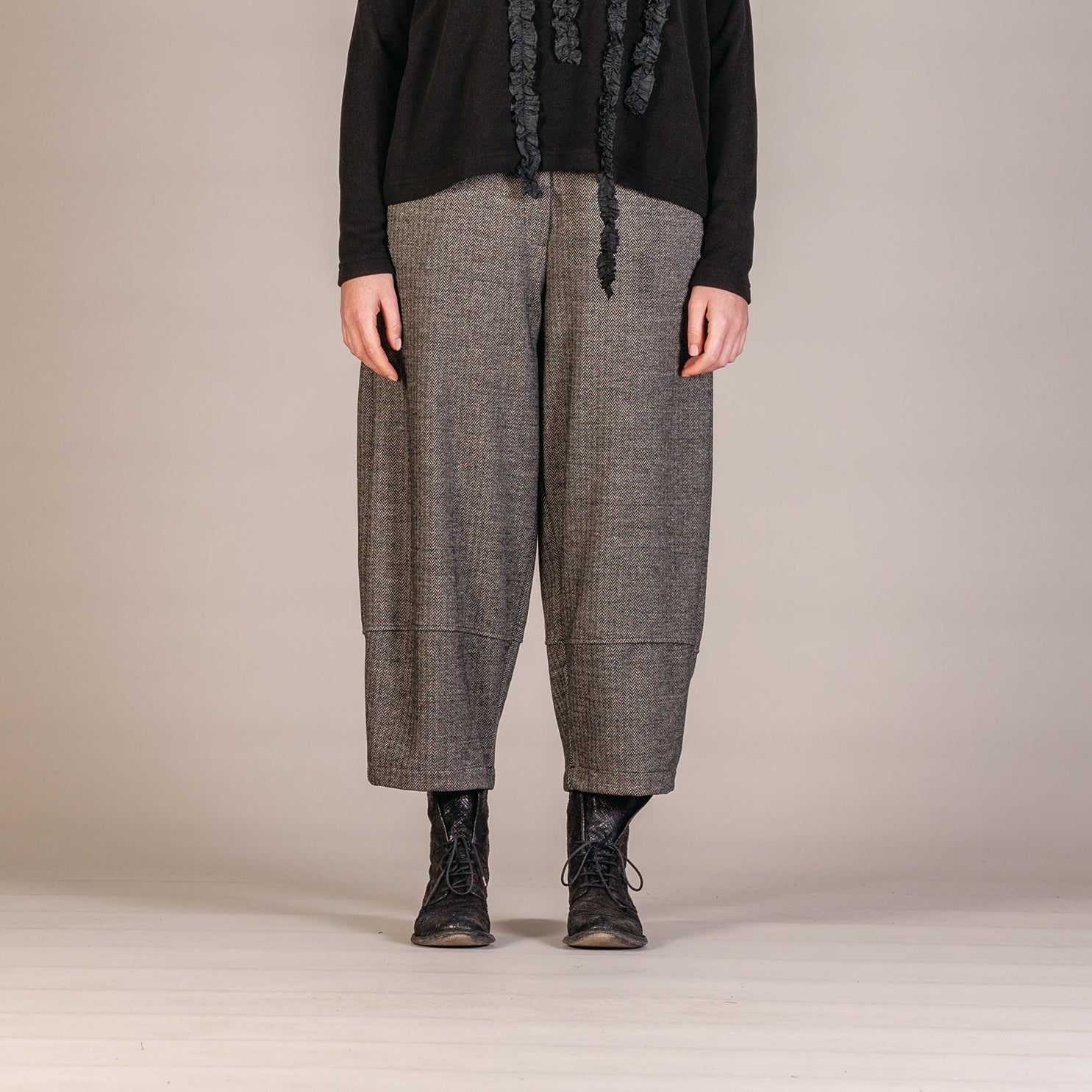 A model stands against a neutral beige studio background wearing a black long-sleeve top with vertical textured fringe details down the front, paired with loose gray wide-leg trousers and black lace-up boots.