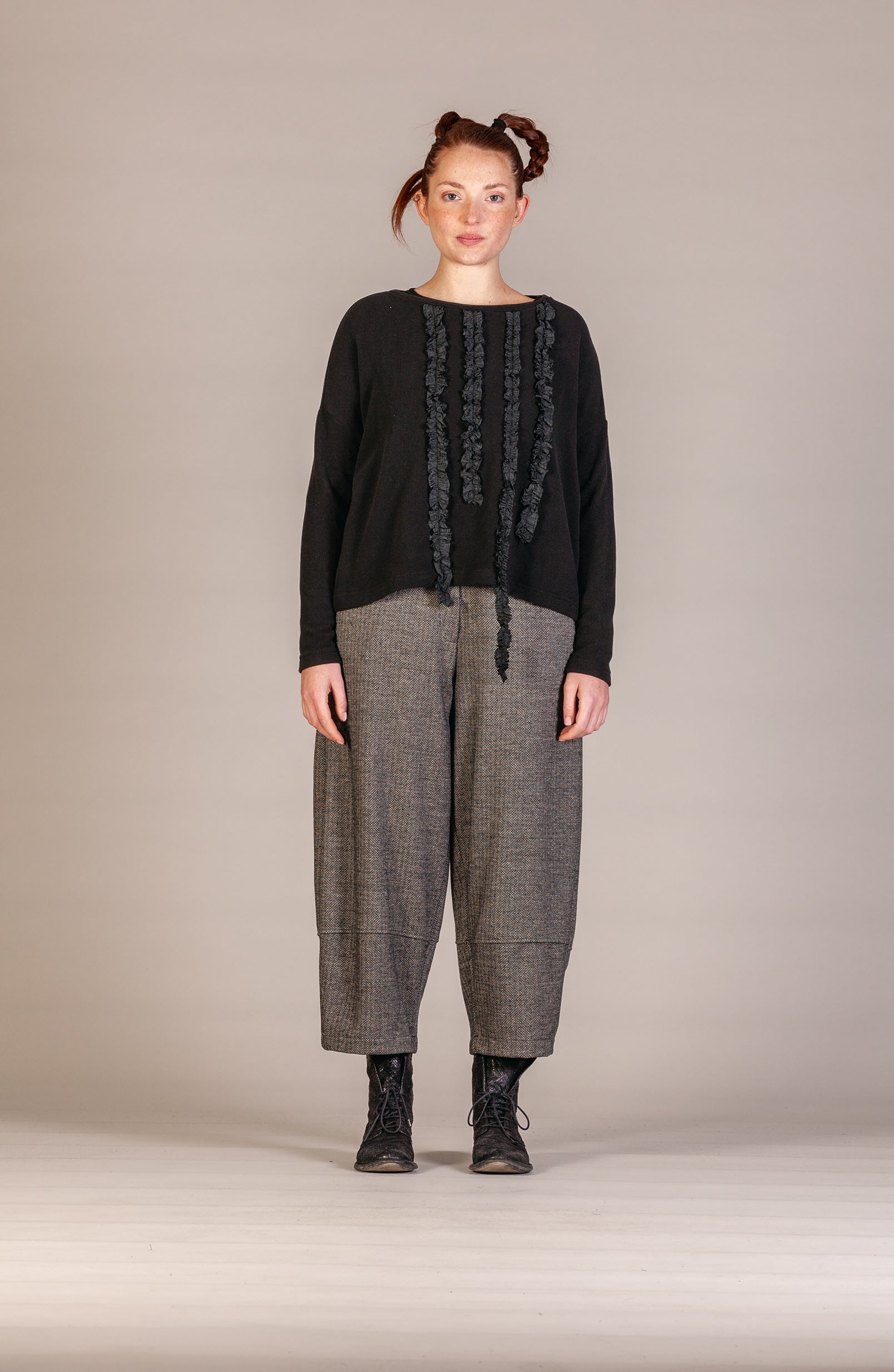 A model stands against a neutral beige studio background wearing a black long-sleeve top with vertical textured fringe details down the front, paired with loose gray wide-leg trousers and black lace-up boots.