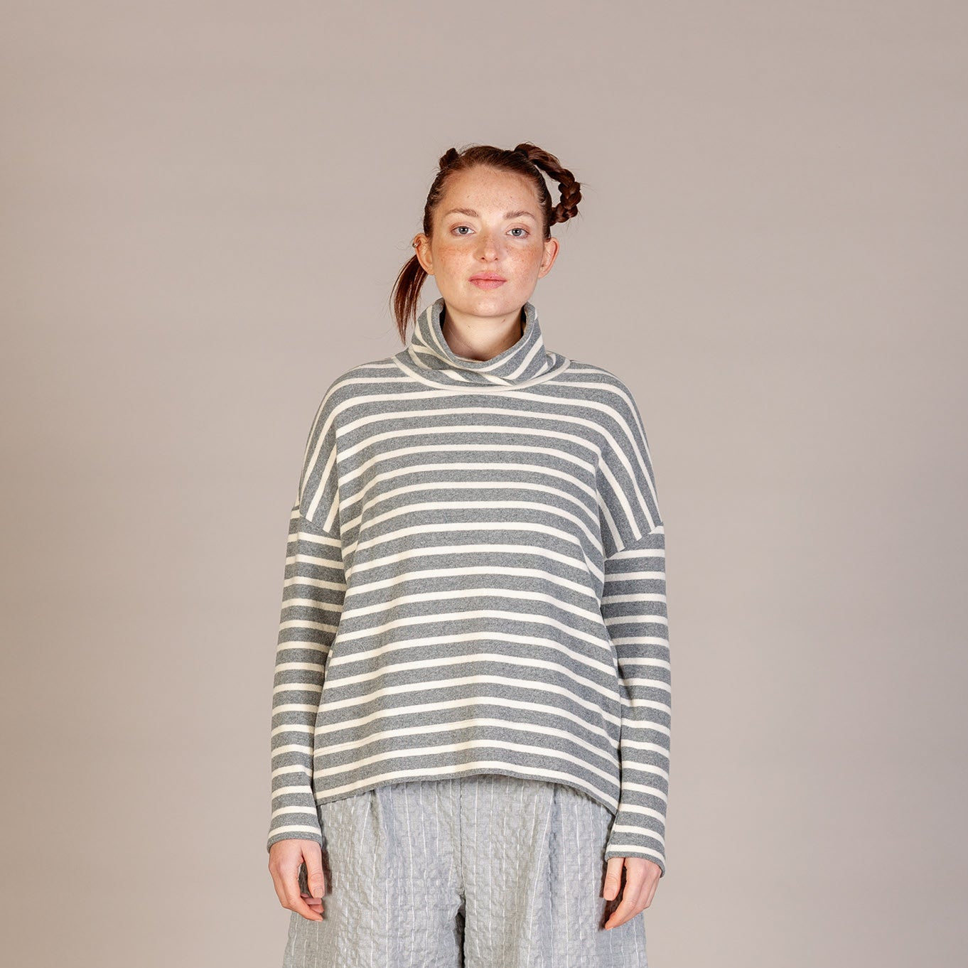A woman stands facing forward in a studio wearing a loose striped turtleneck sweater and loose light gray pants with dark lace-up boots. The background is a plain warm light gray/beige studio backdrop.