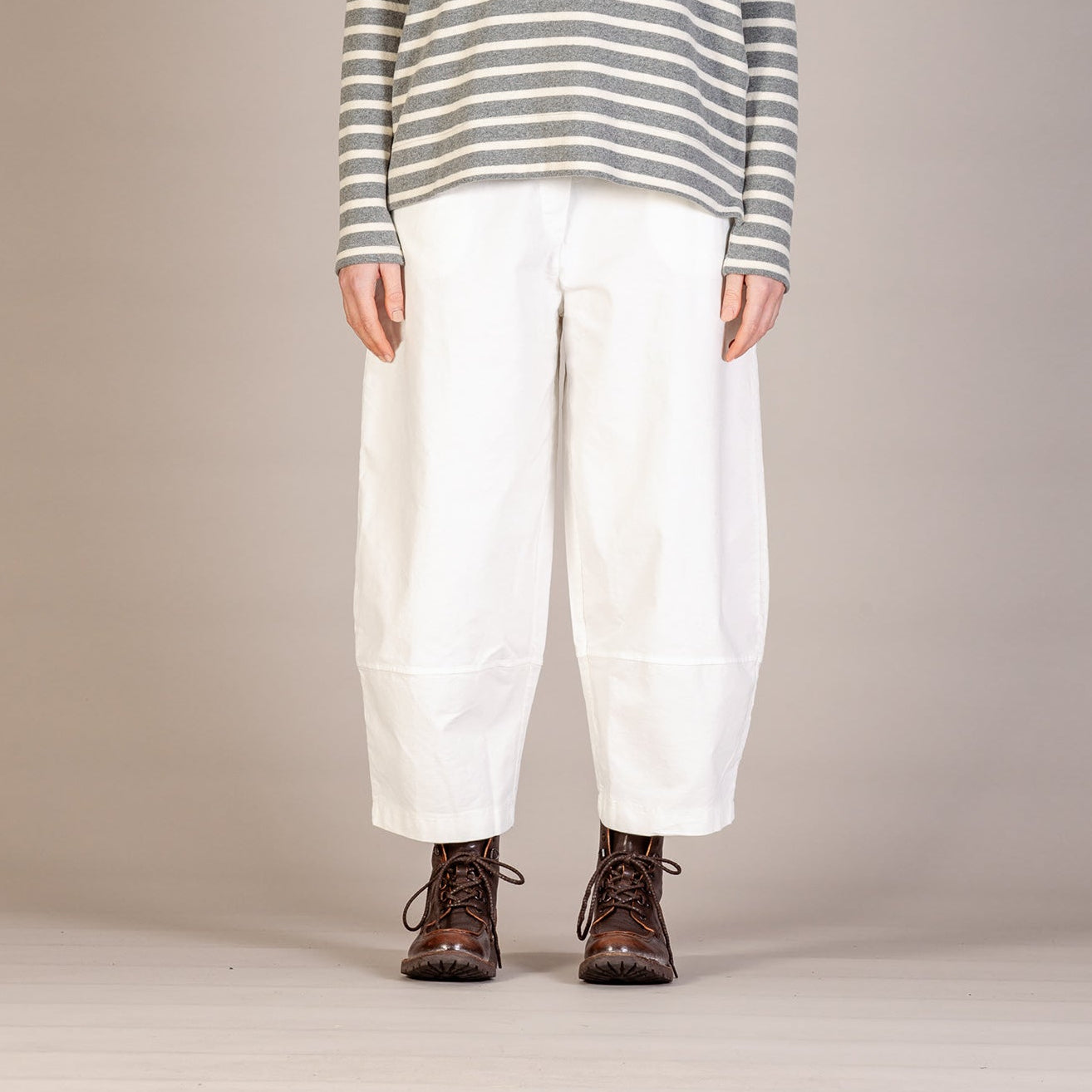 A model stands against a neutral beige studio background wearing a gray and white horizontally striped turtleneck sweater, loose white pants, and dark brown lace-up boots.