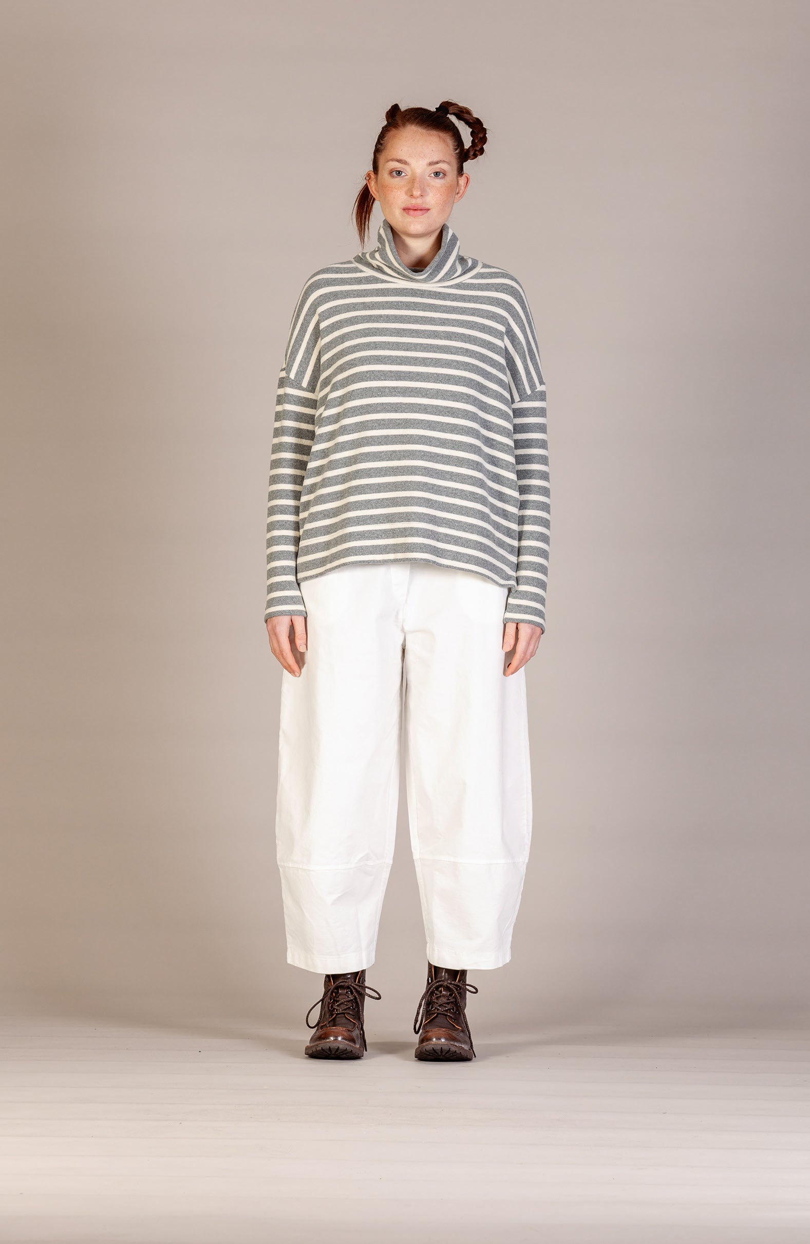 A model stands against a neutral beige studio background wearing a gray and white horizontally striped turtleneck sweater, loose white pants, and dark brown lace-up boots.