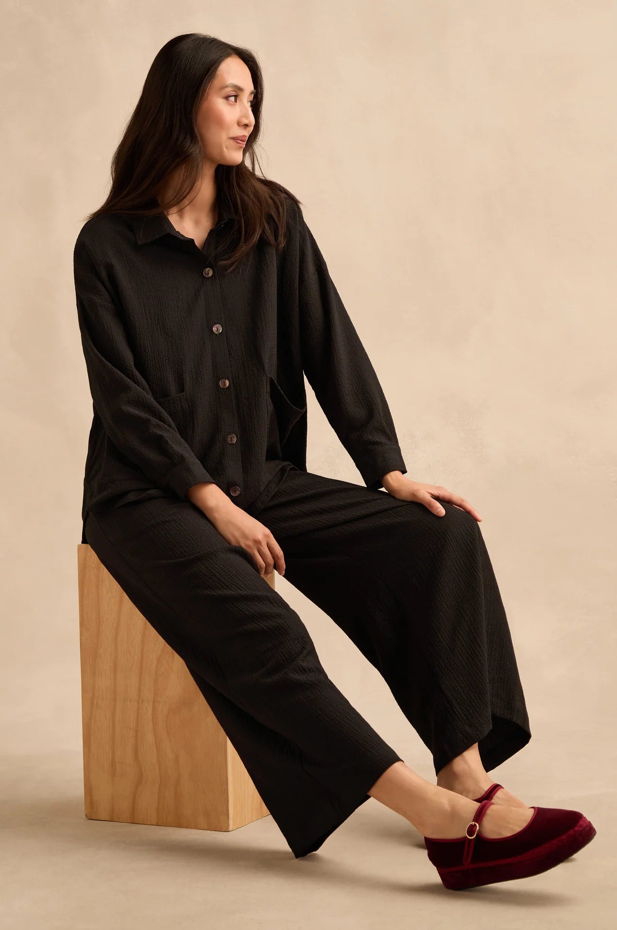 A woman sits on a wooden block wearing a black long-sleeve button-front top and matching black wide-leg pants, paired with burgundy Mary Jane shoes, against a warm beige studio background.