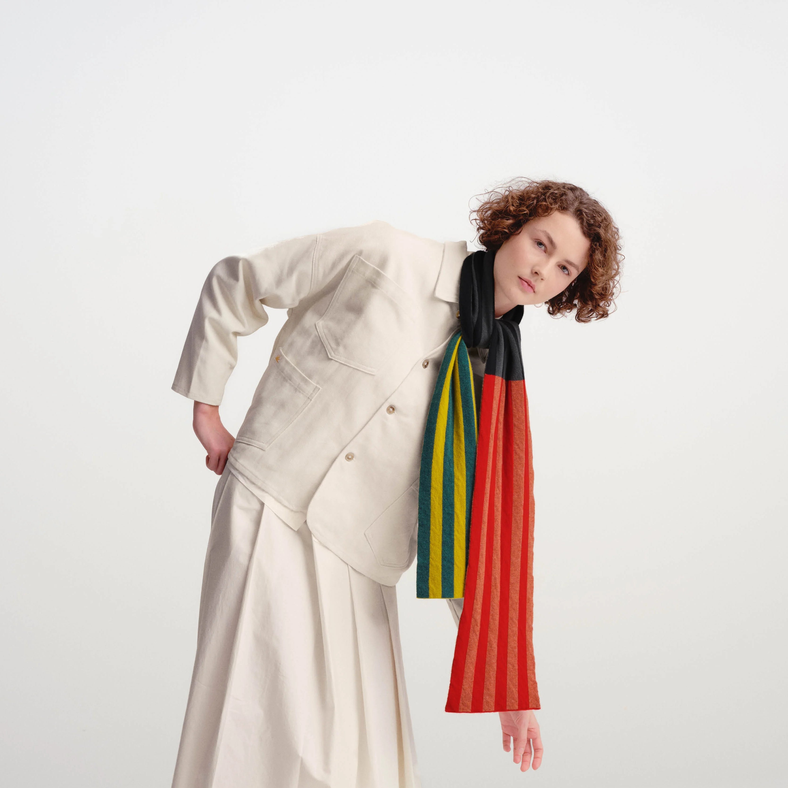 A model wearing a cream-colored asymmetrical jacket and matching midi skirt, styled with a vibrant red, green, yellow, and black striped knit scarf, cream tights, and white leather shoes, set against a plain light gray studio background.