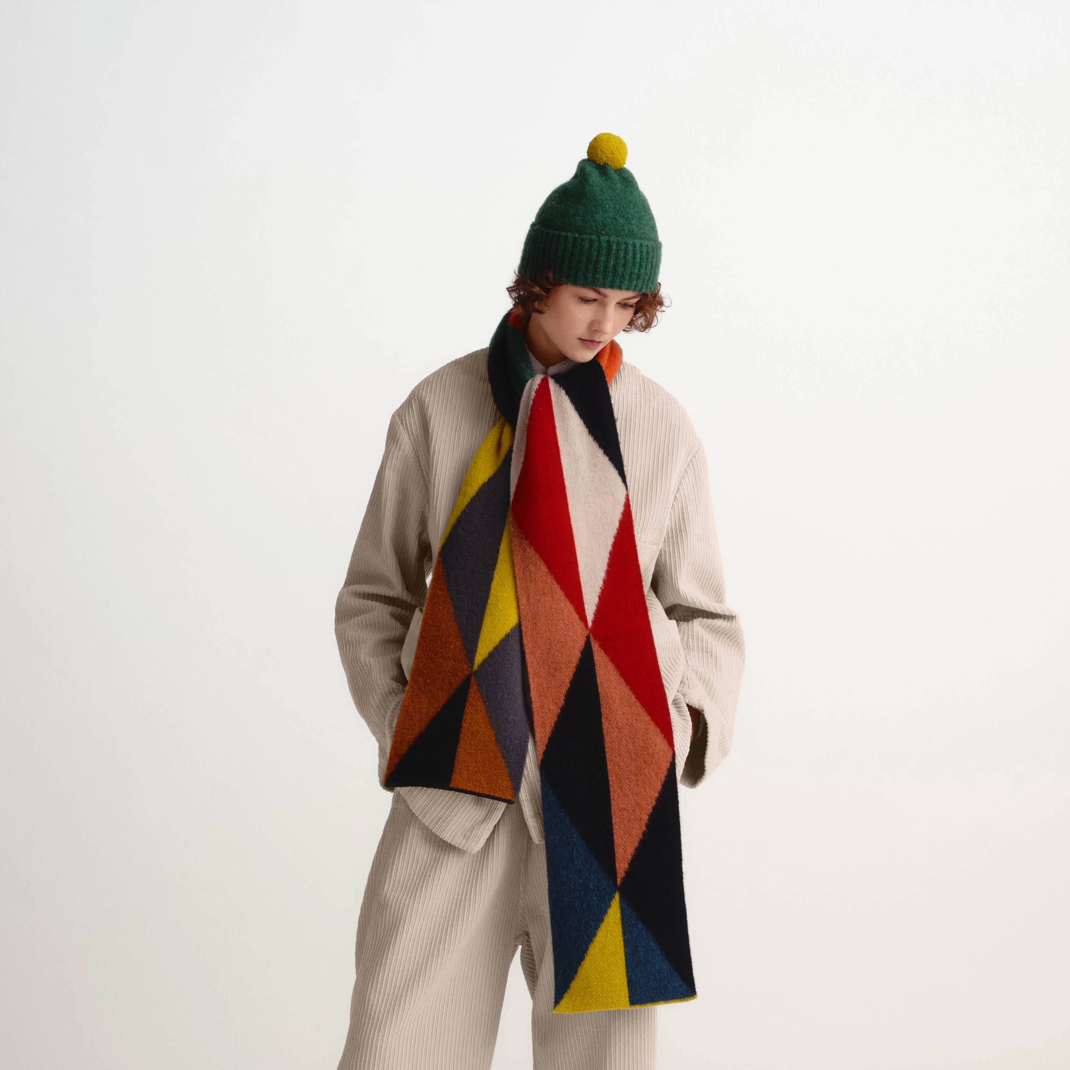 A person stands in a studio wearing a beige coat and matching wide-leg pants, with a large multicolor geometric scarf draped around the neck. The person also wears a green knit beanie with a yellow pom-pom and white shoes.