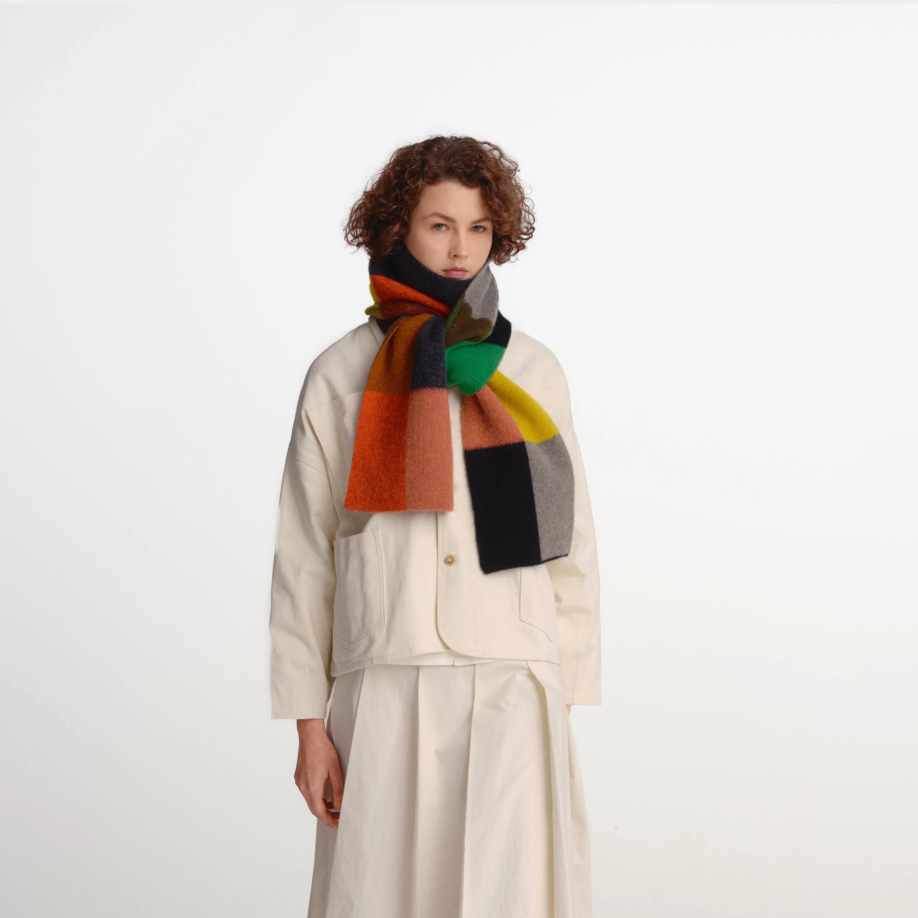 A model wearing a cream-colored layered jacket and skirt set with a vibrant multicolor geometric scarf, standing against a plain light gray studio background.