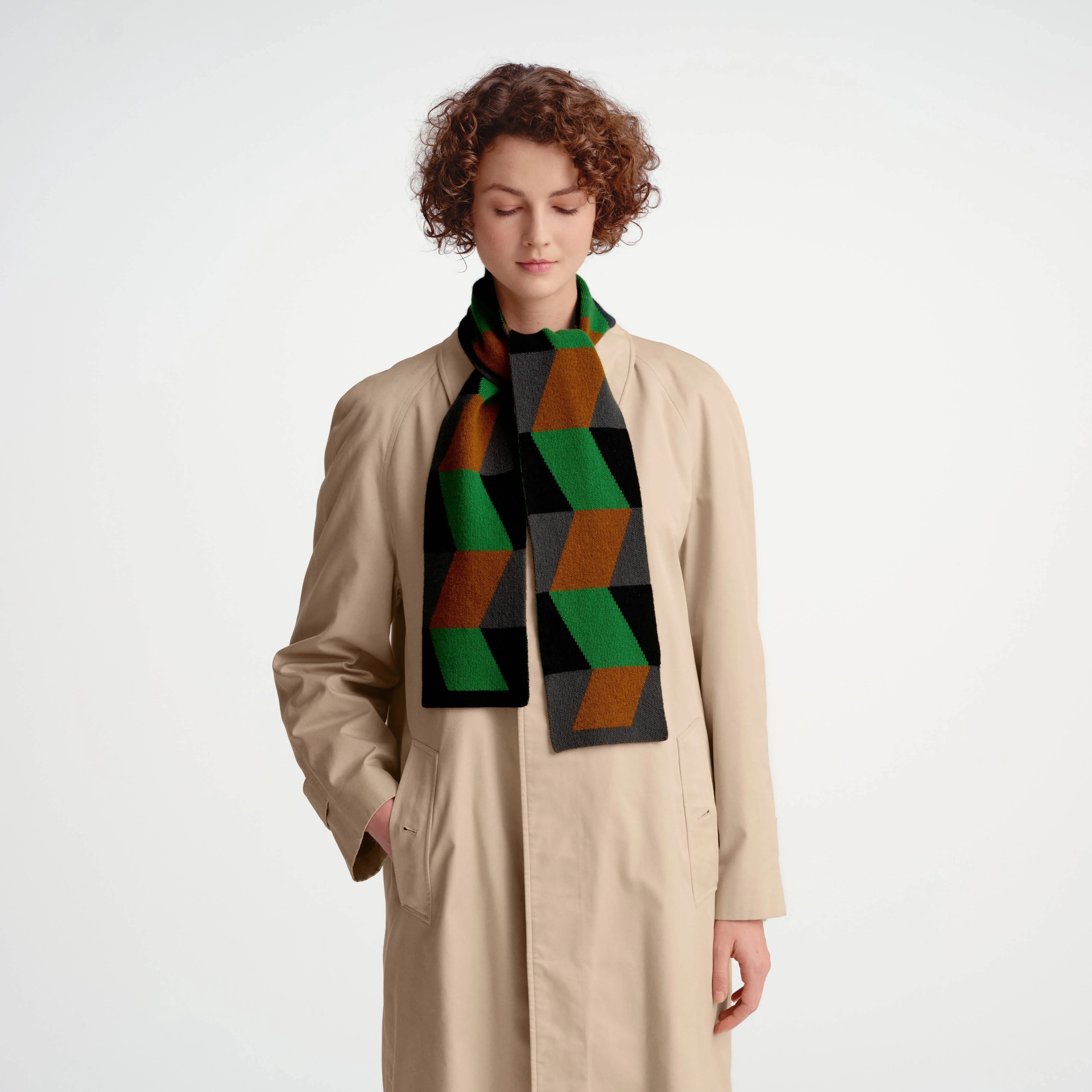A model wearing a beige trench coat and a green, black, and brown geometric patterned knit scarf, standing against a plain light gray studio background.