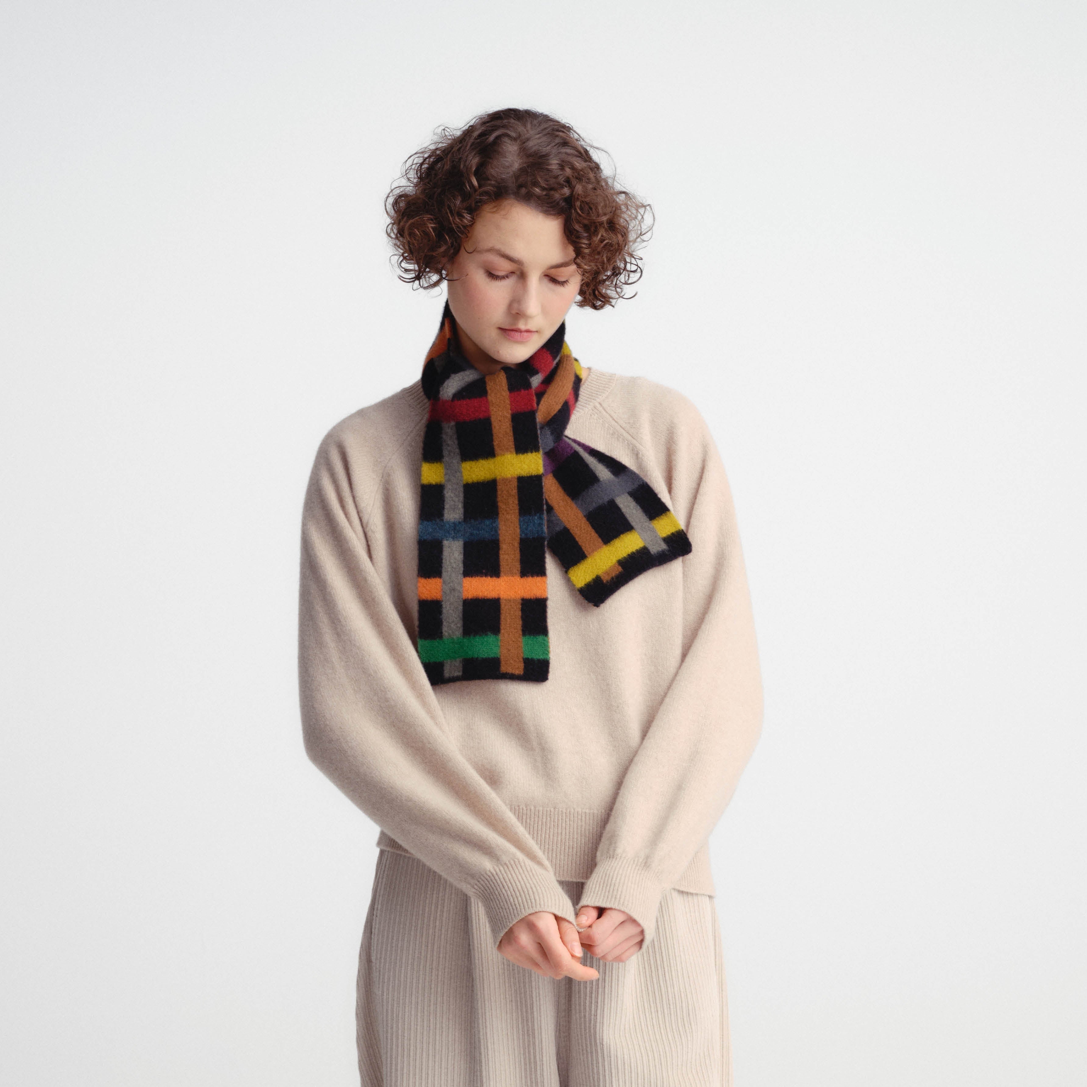 A model wearing a beige ribbed knit jumpsuit and a colorful plaid scarf in black, yellow, green, orange, and red, standing against a light gray studio background.