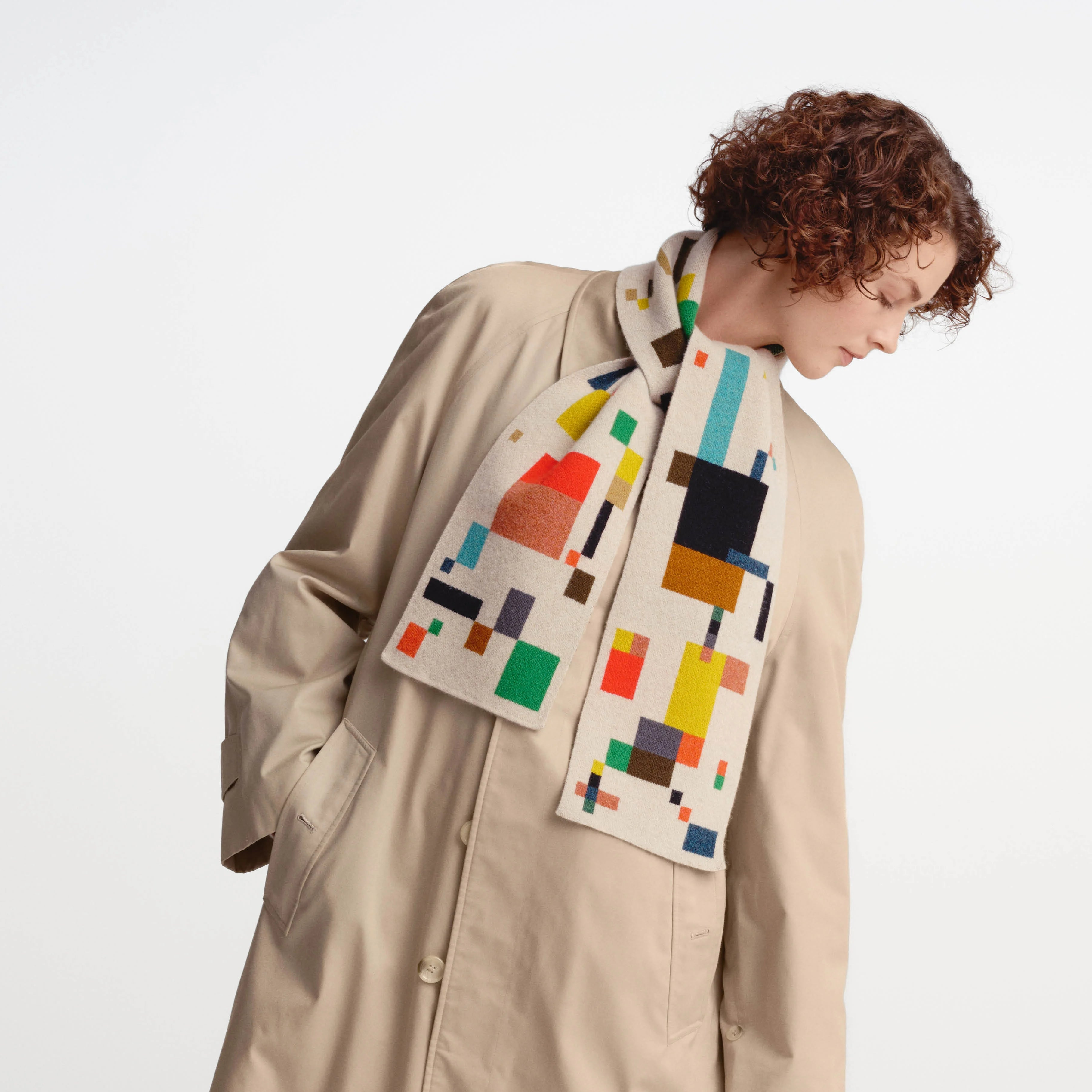 A model wearing a beige trench coat and a cream-colored knit scarf with a colorful geometric block pattern, photographed against a plain white studio background.