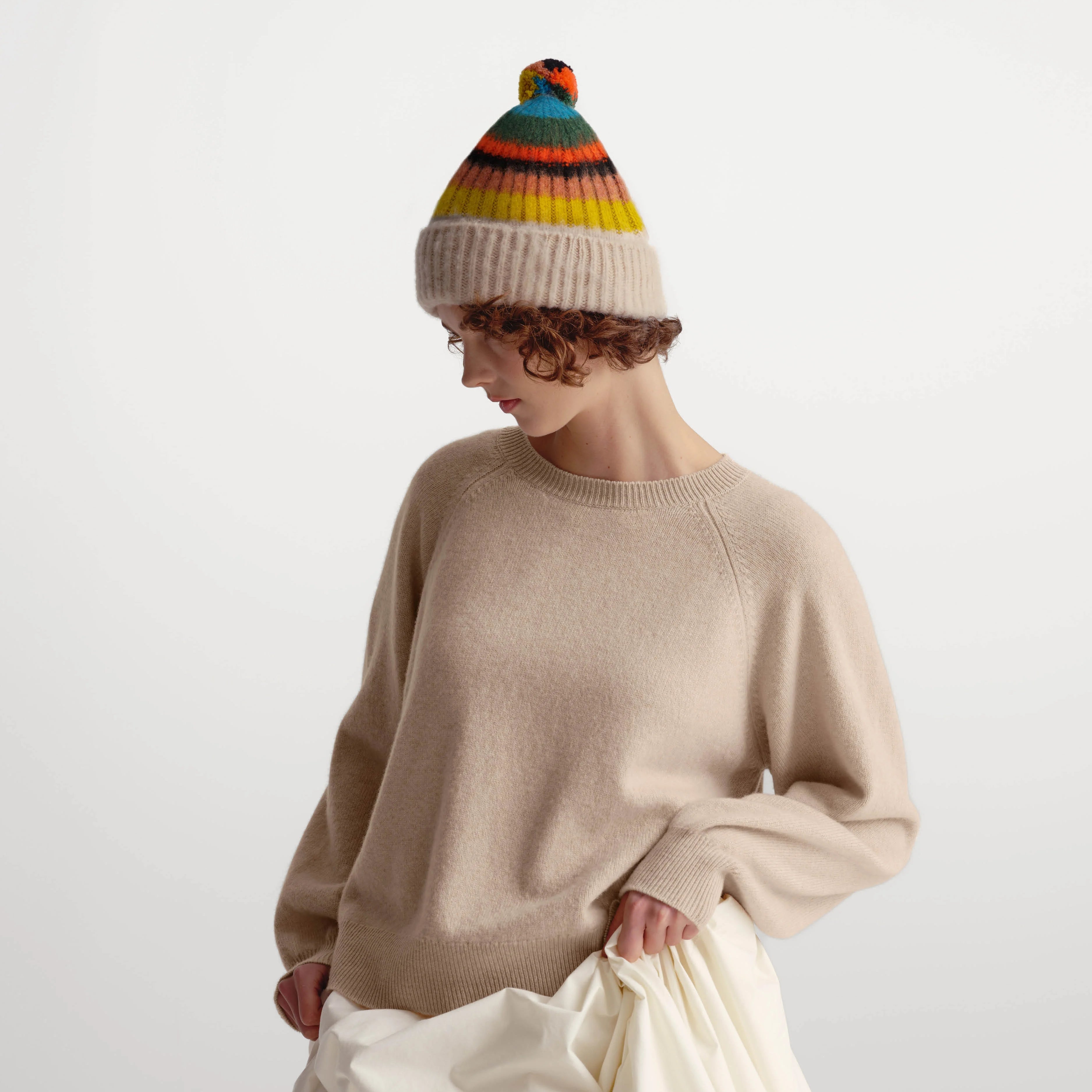 A model wearing a beige knit crewneck sweater, a cream pleated skirt, and a colorful striped beanie with a pom-pom, photographed against a light gray studio background.