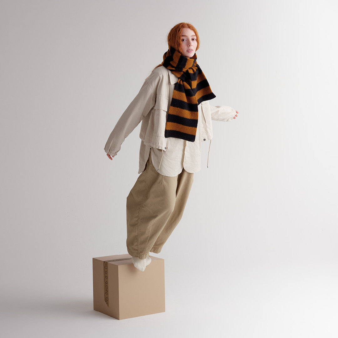 Woman standing on a box wearing a beige shirt, khaki pants, and a brown and black striped scarf.