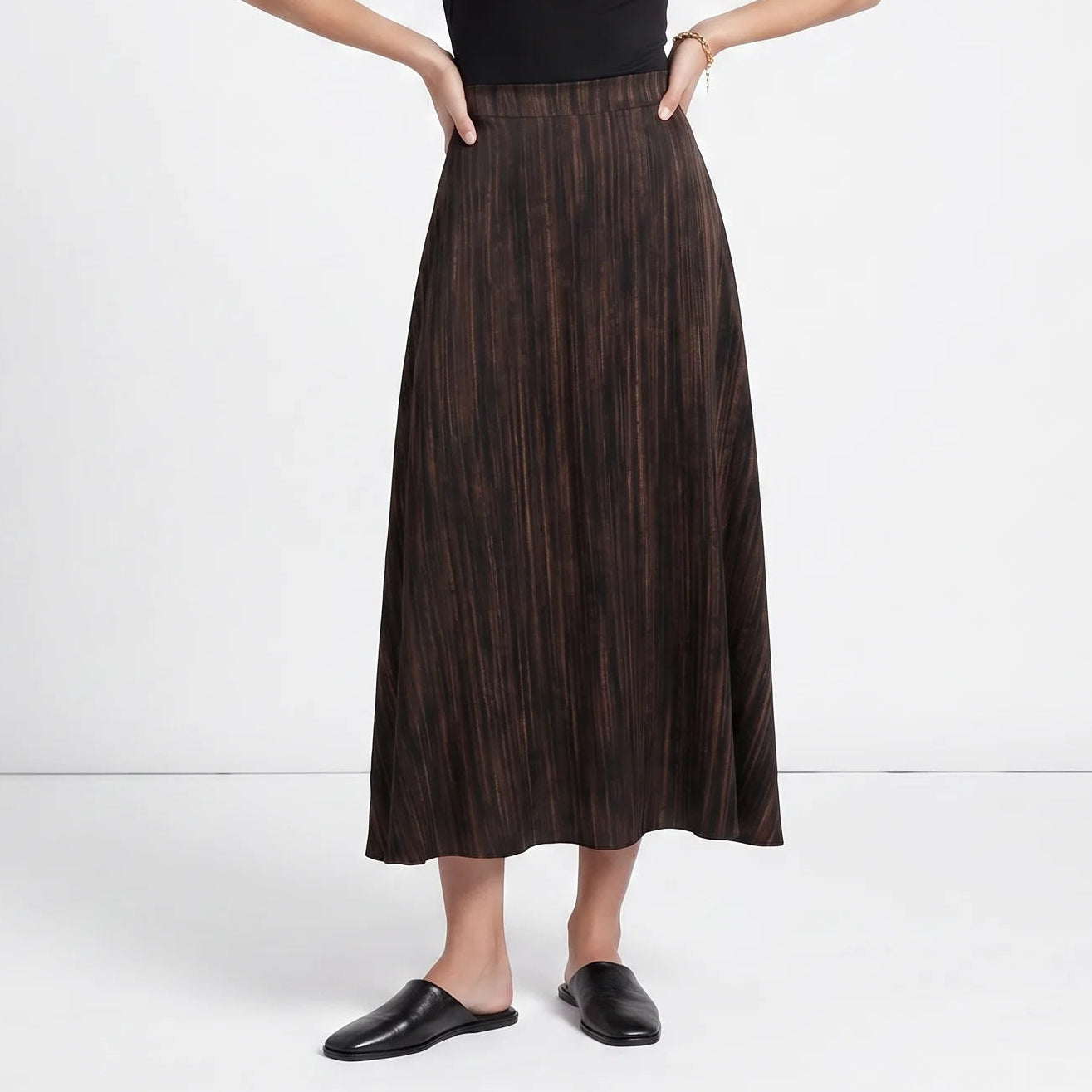 A woman stands in a studio wearing a sleeveless black mock-neck top and a dark brown pleated midi skirt, paired with black loafers.