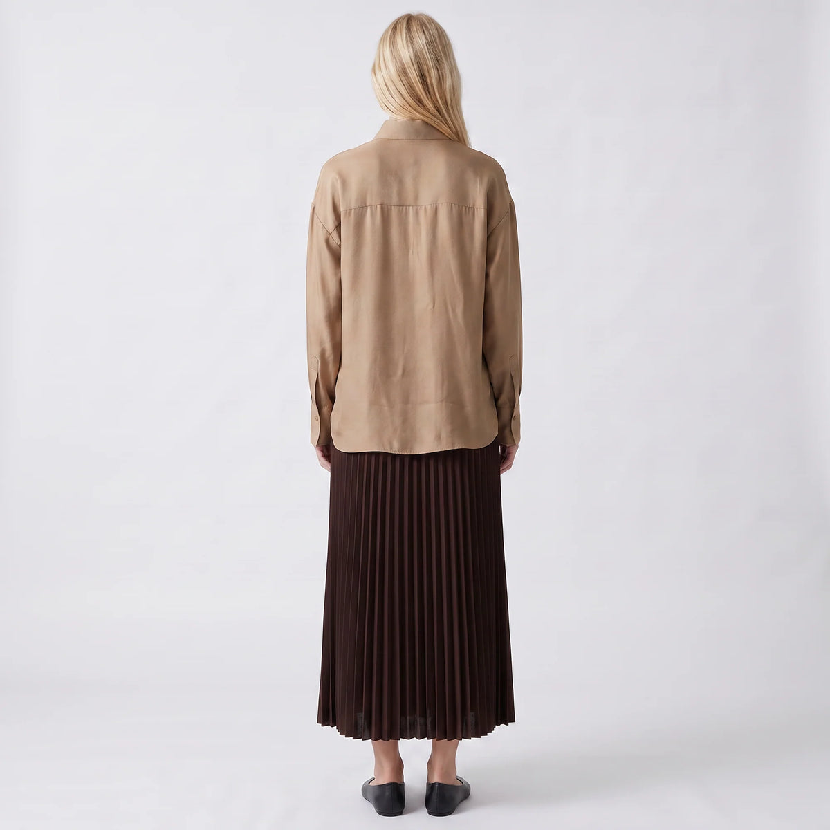 A woman stands with her back to the camera wearing a light brown long-sleeve shirt and a dark brown pleated midi skirt, set against a plain white studio background.