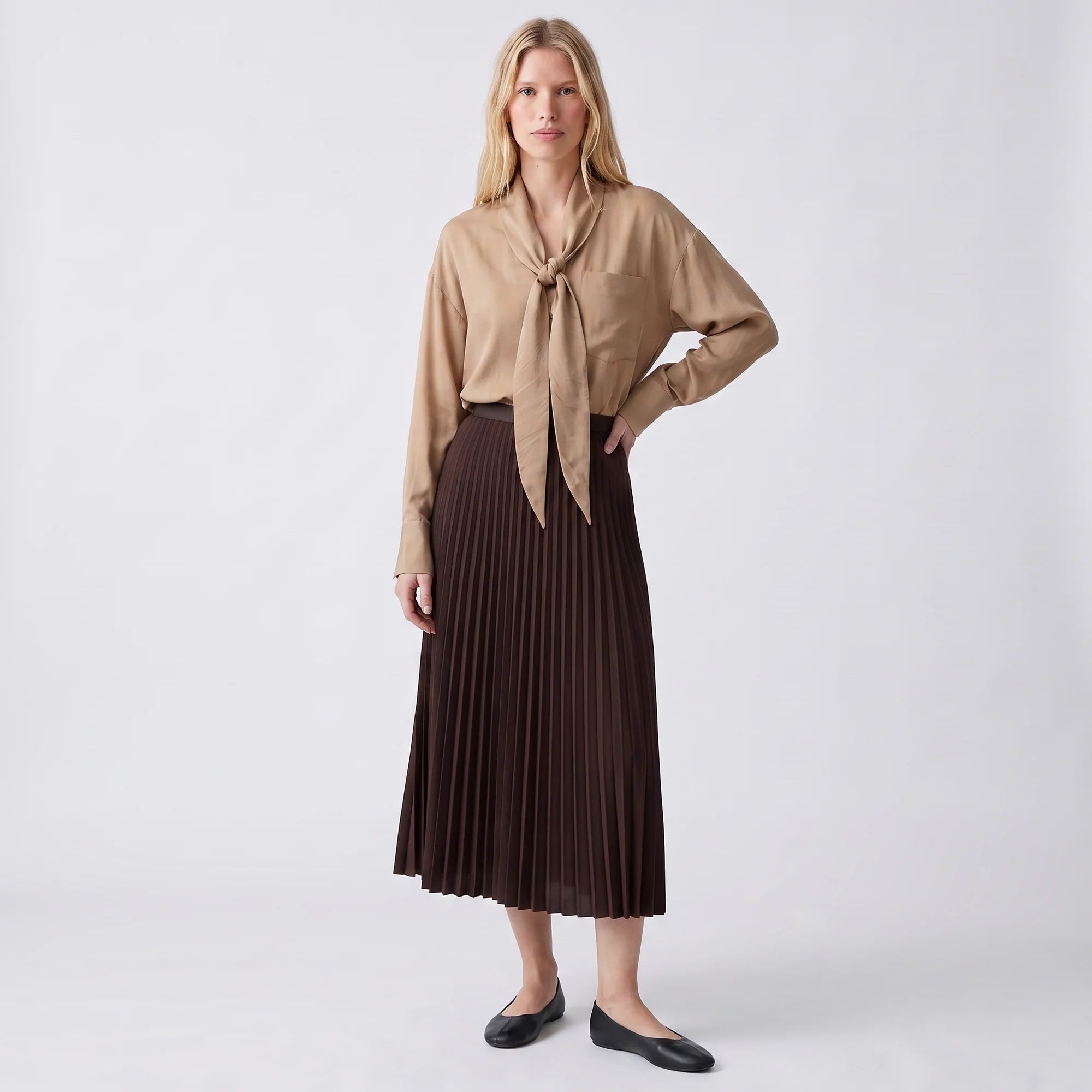 A woman modeling a beige long-sleeve blouse with a front tie knot and a dark brown pleated midi skirt, paired with black flats, against a light gray studio background.