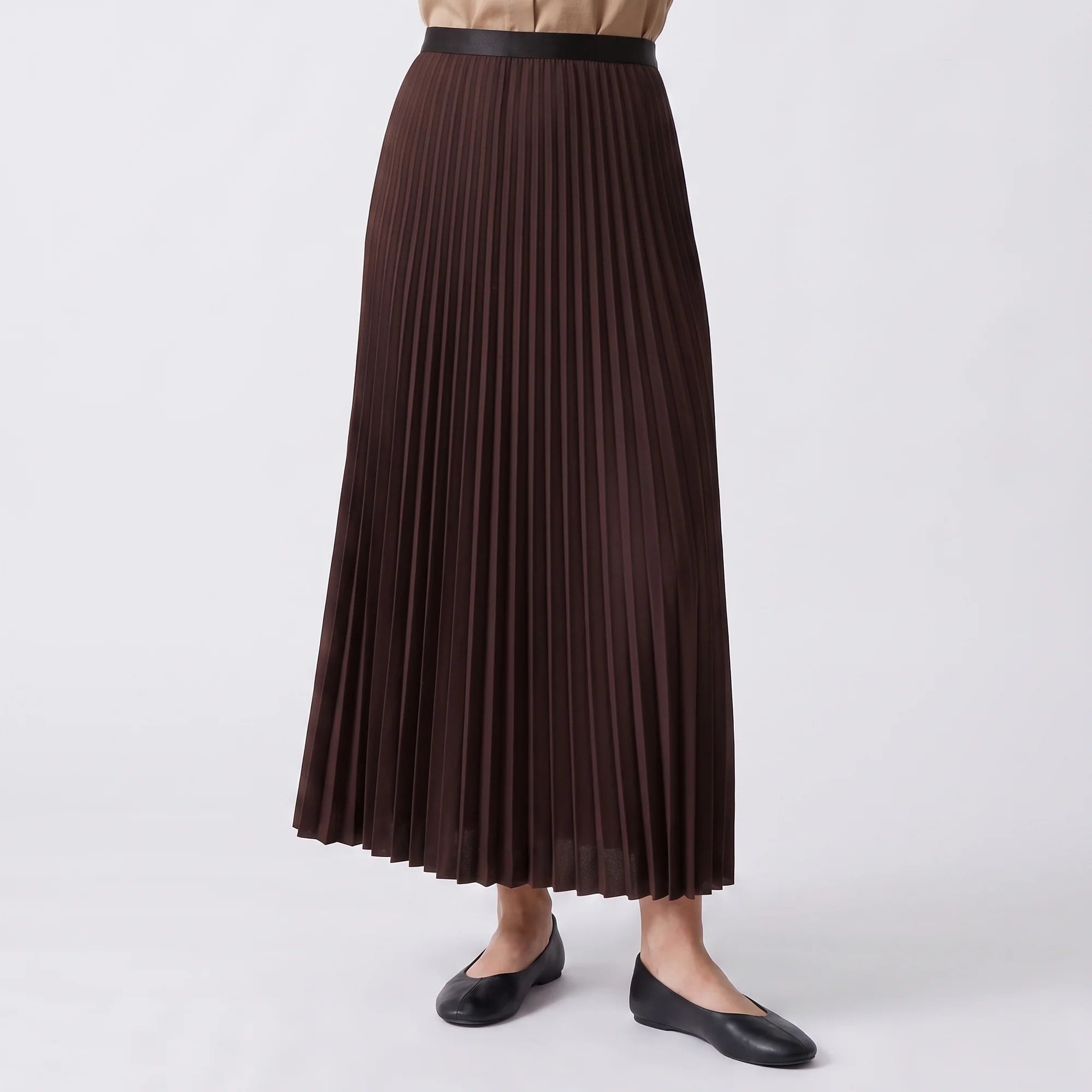 A dark brown pleated midi skirt with a black waistband, shown on a model wearing black flats against a light gray studio background.