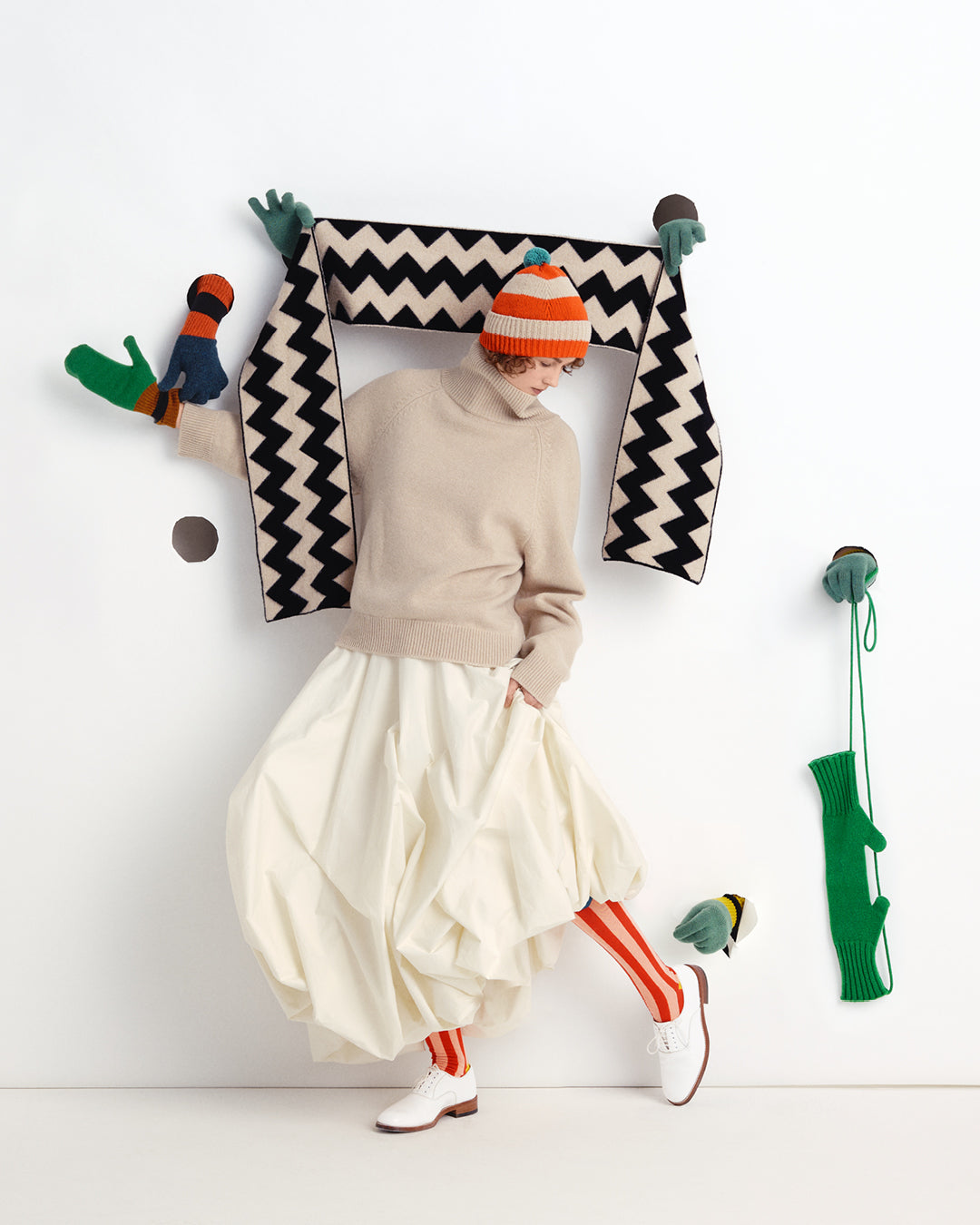 A model wearing a beige turtleneck sweater, a voluminous cream pleated skirt, a striped orange and white beanie, and red-and-white striped socks, posing with a black and white chevron knit scarf and colorful knit accessories against a white wall.