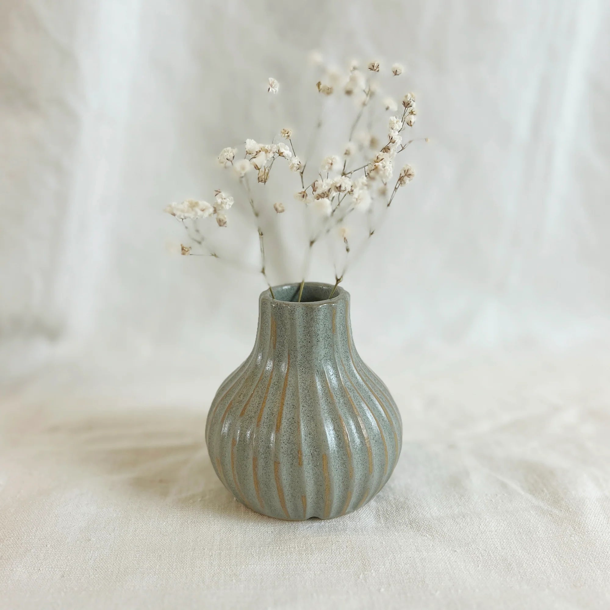 Small, ribbed, gray ceramic vase with a bulbous shape, containing a sprig of dried white flowers.
