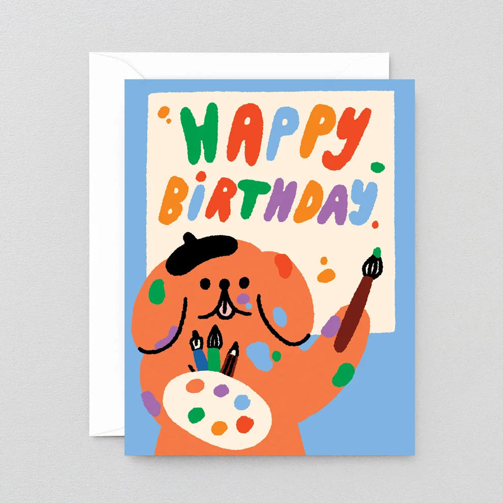 A colorful 'Happy Birthday' greeting card featuring a cartoon dog painted like an artist, holding a paint palette and brush, with a blue border and white envelope behind it, shown on a light gray background.