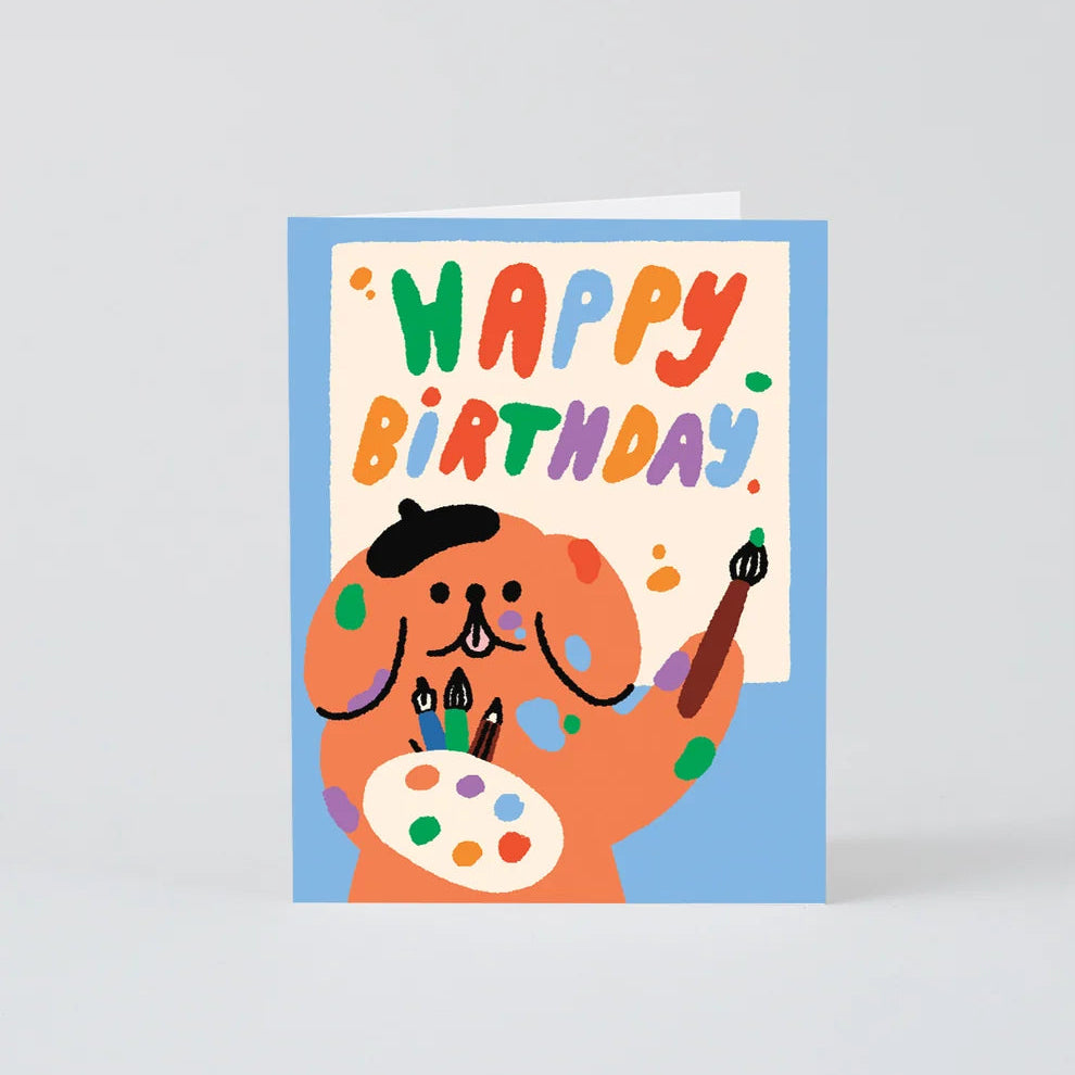 A square birthday card featuring a cartoon dog painting with colorful paint splatters and the words “HAPPY BIRTHDAY” in multicolored letters, shown on a light gray background.