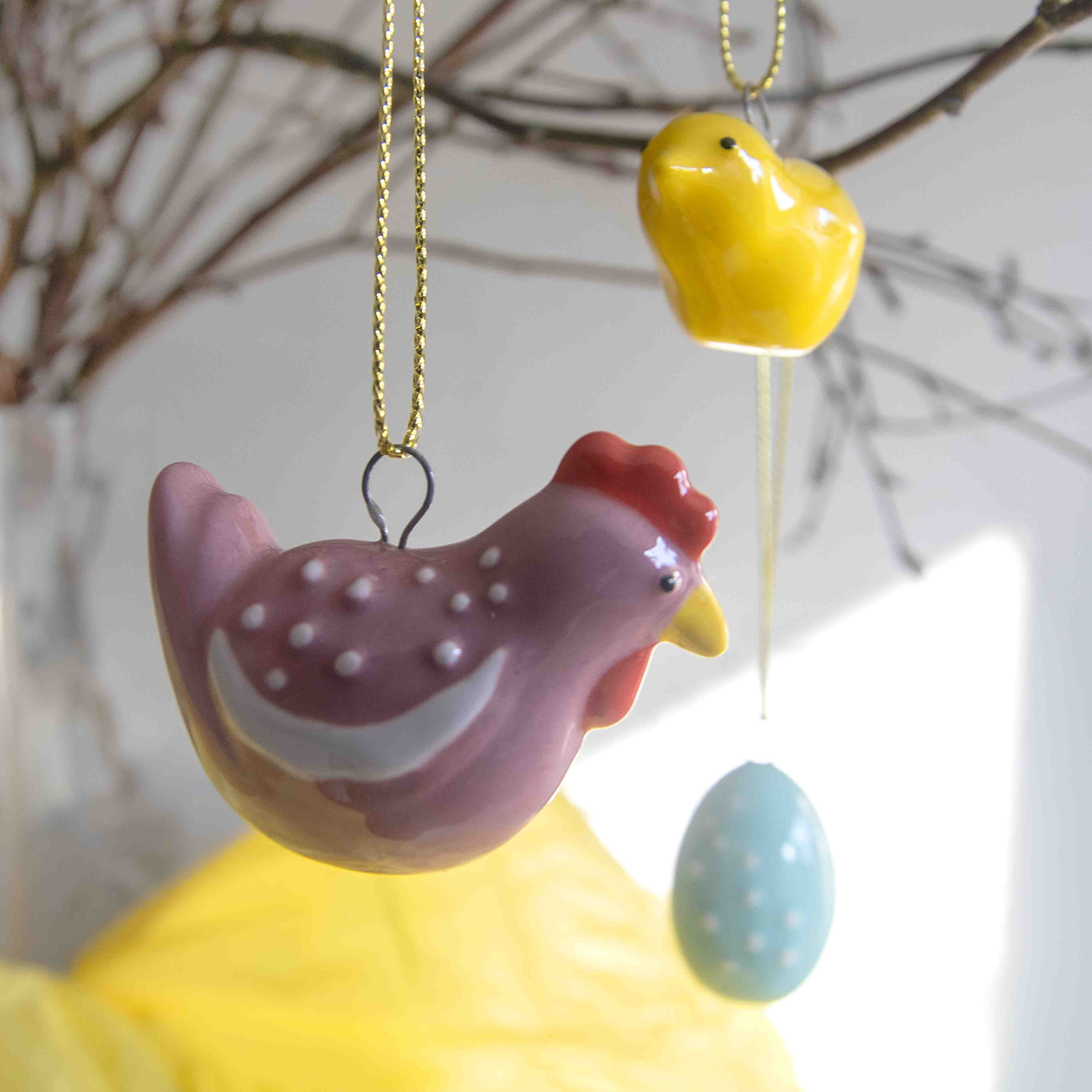 Three ceramic Easter ornaments: a pink hen with white dots, a yellow chick, and a light blue egg, each hanging from a gold