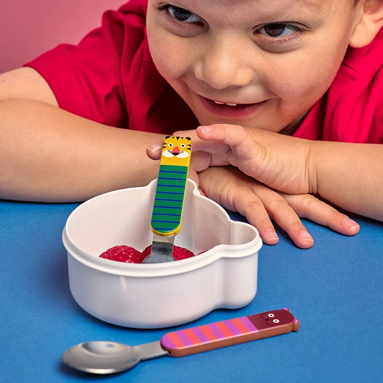 Child using a colorful children's fork and spoon set with a bowl of fruit on a blue surface.