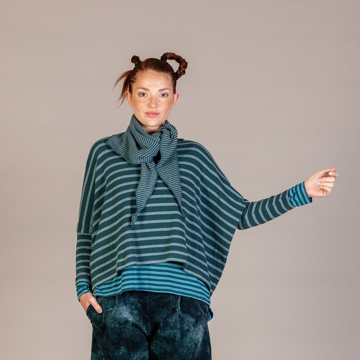 A model stands against a neutral studio backdrop wearing a teal and dark green striped knit top with a matching scarf, paired with dark blue-green tie-dye pants and brown shoes.