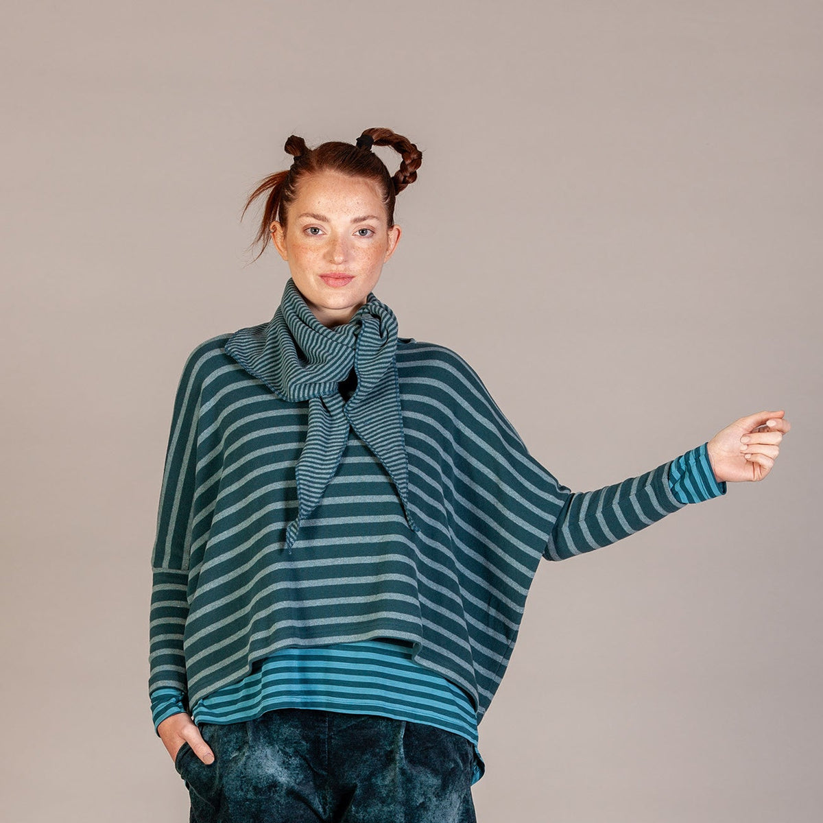 A model stands against a neutral studio backdrop wearing a teal and dark green striped knit top with a matching scarf, paired with dark blue-green tie-dye pants and brown shoes.