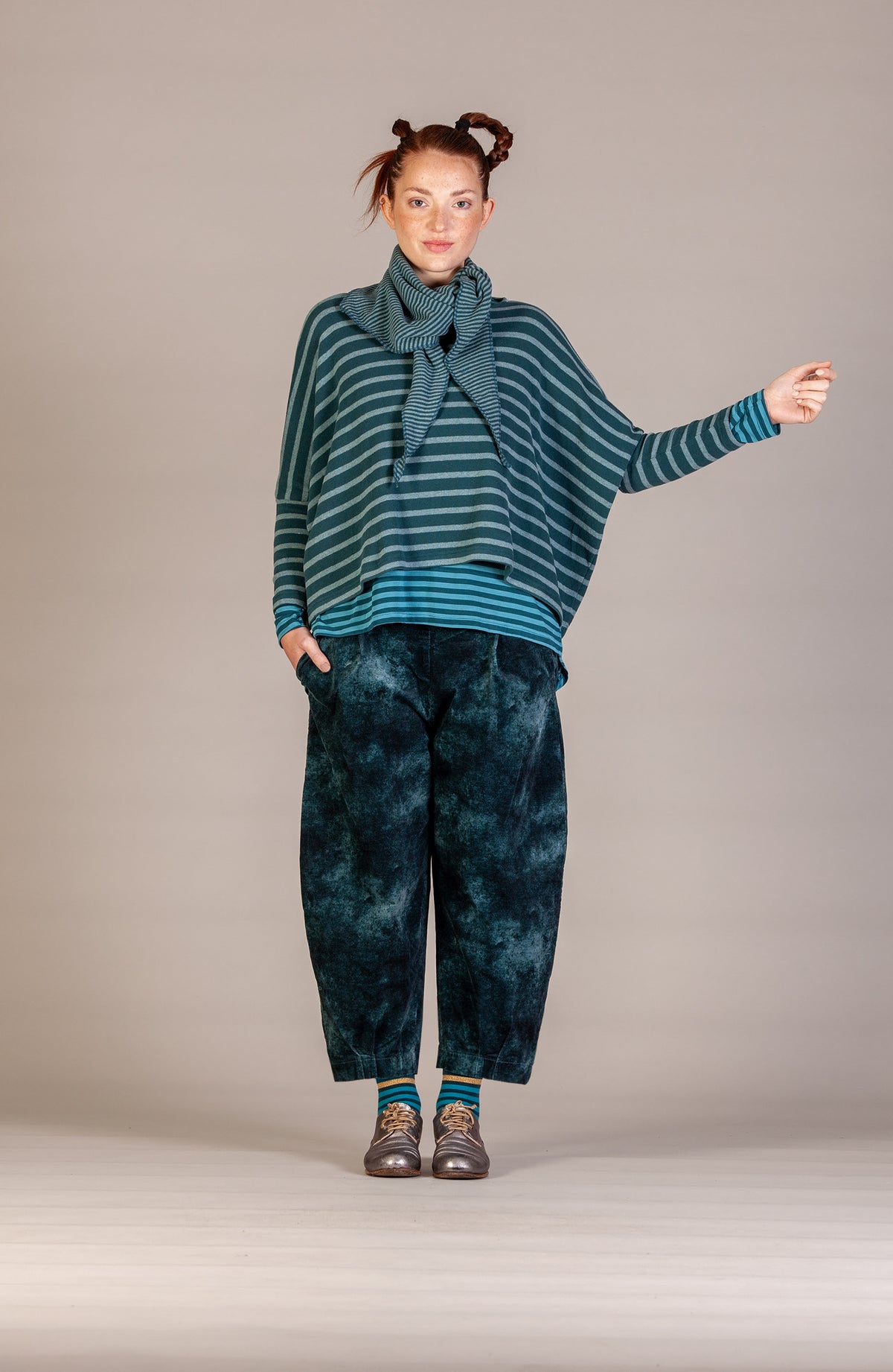 A model stands against a neutral studio backdrop wearing a teal and dark green striped knit top with a matching scarf, paired with dark blue-green tie-dye pants and brown shoes.
