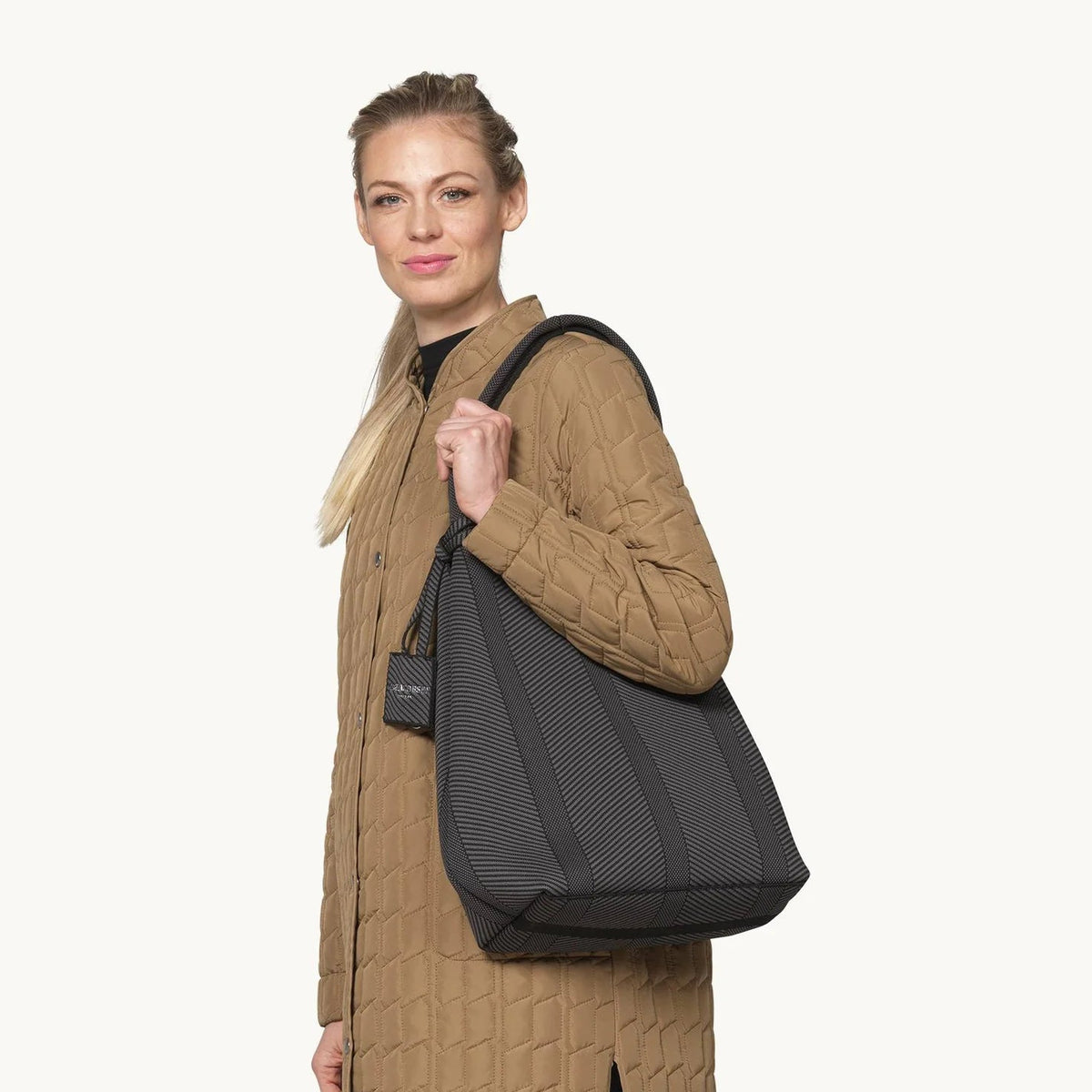 A woman modeling a large black tote bag with a shoulder strap, styled with a brown quilted coat, blue jeans, and brown shoes against a plain off-white background.