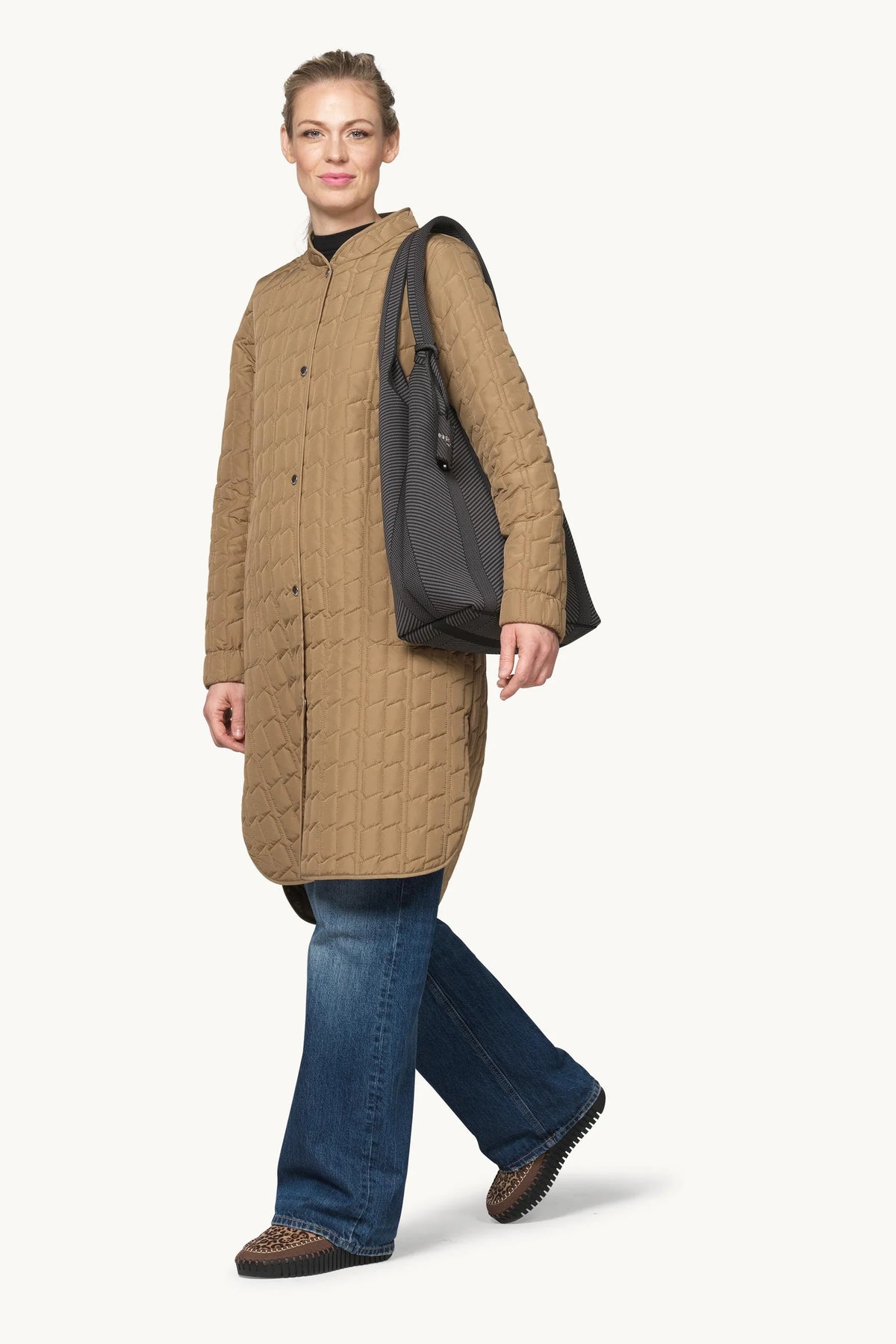 A woman modeling a long quilted camel-brown coat with a stand collar, paired with blue jeans and a black shoulder bag, against a plain off-white background.