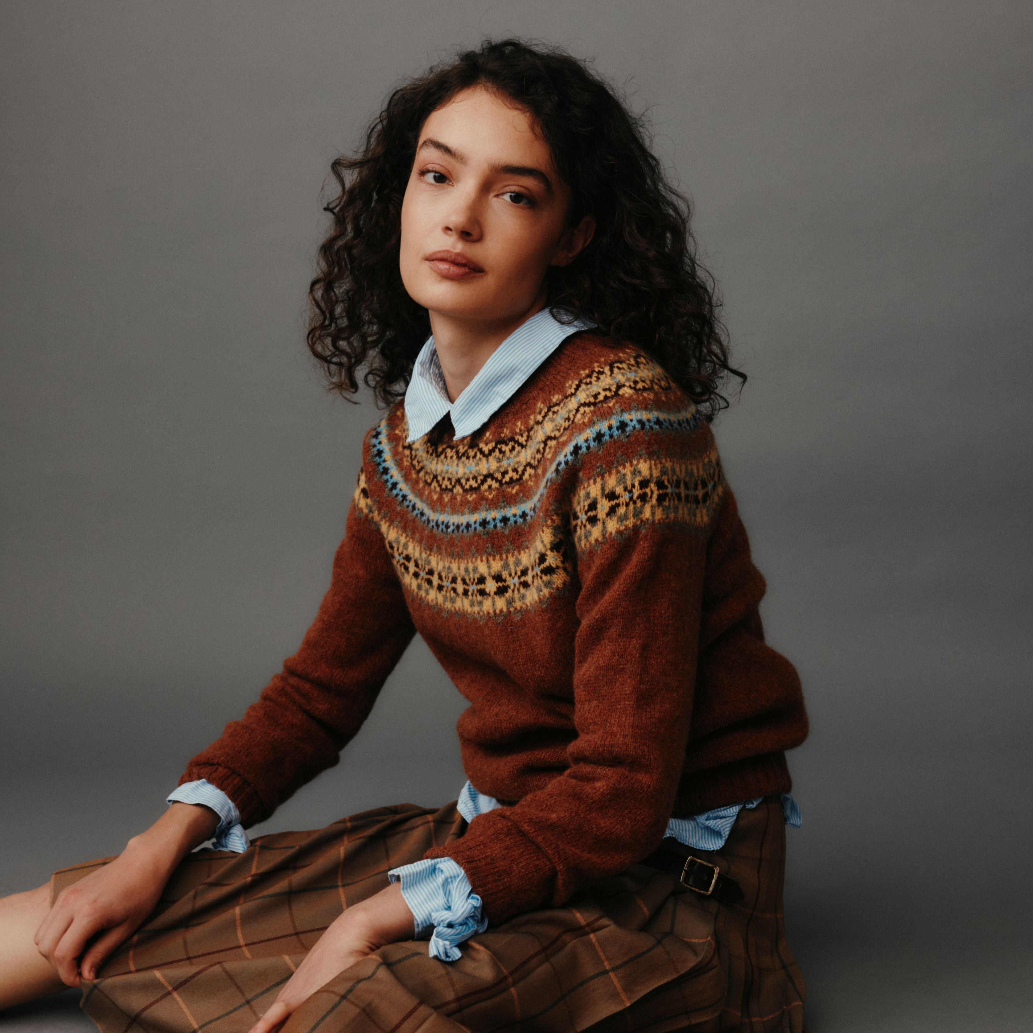 Woman wearing a patterned brown sweater sitting against a gray background