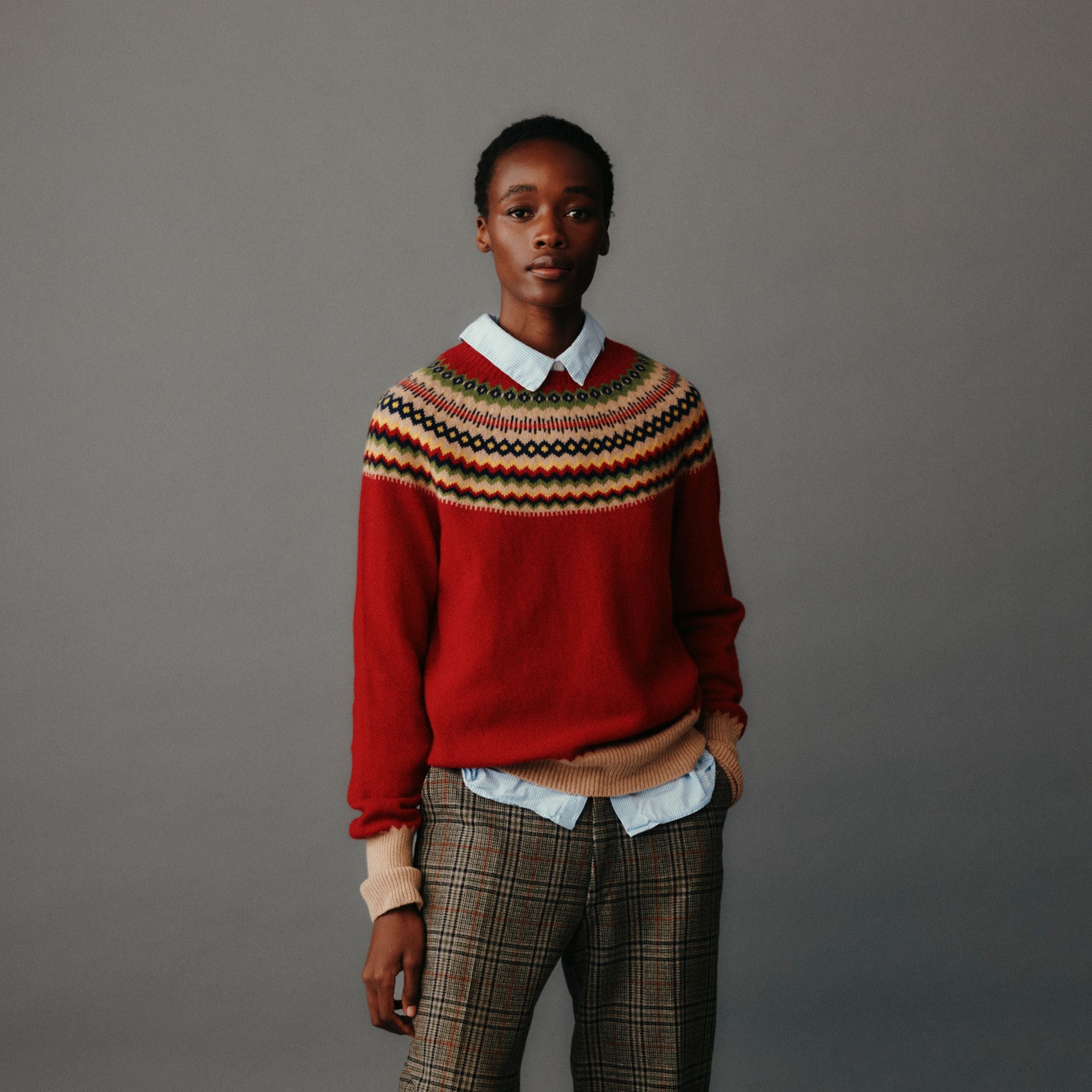 Person wearing a red sweater with a patterned design, plaid pants, and white socks on a gray background