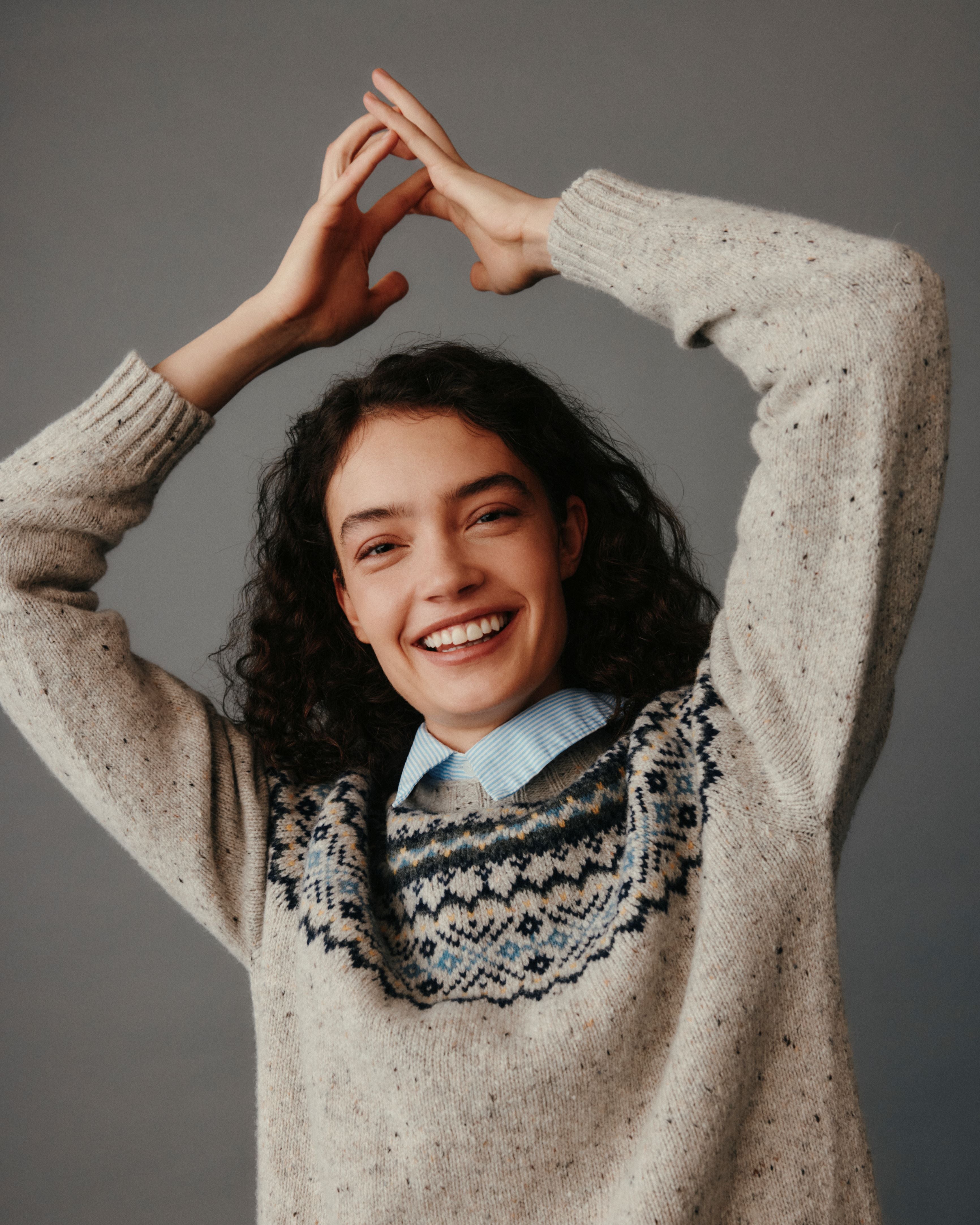 Woman wearing a beige sweater with a patterned neckline, smiling and making a heart shape with her hands against a gray background.
