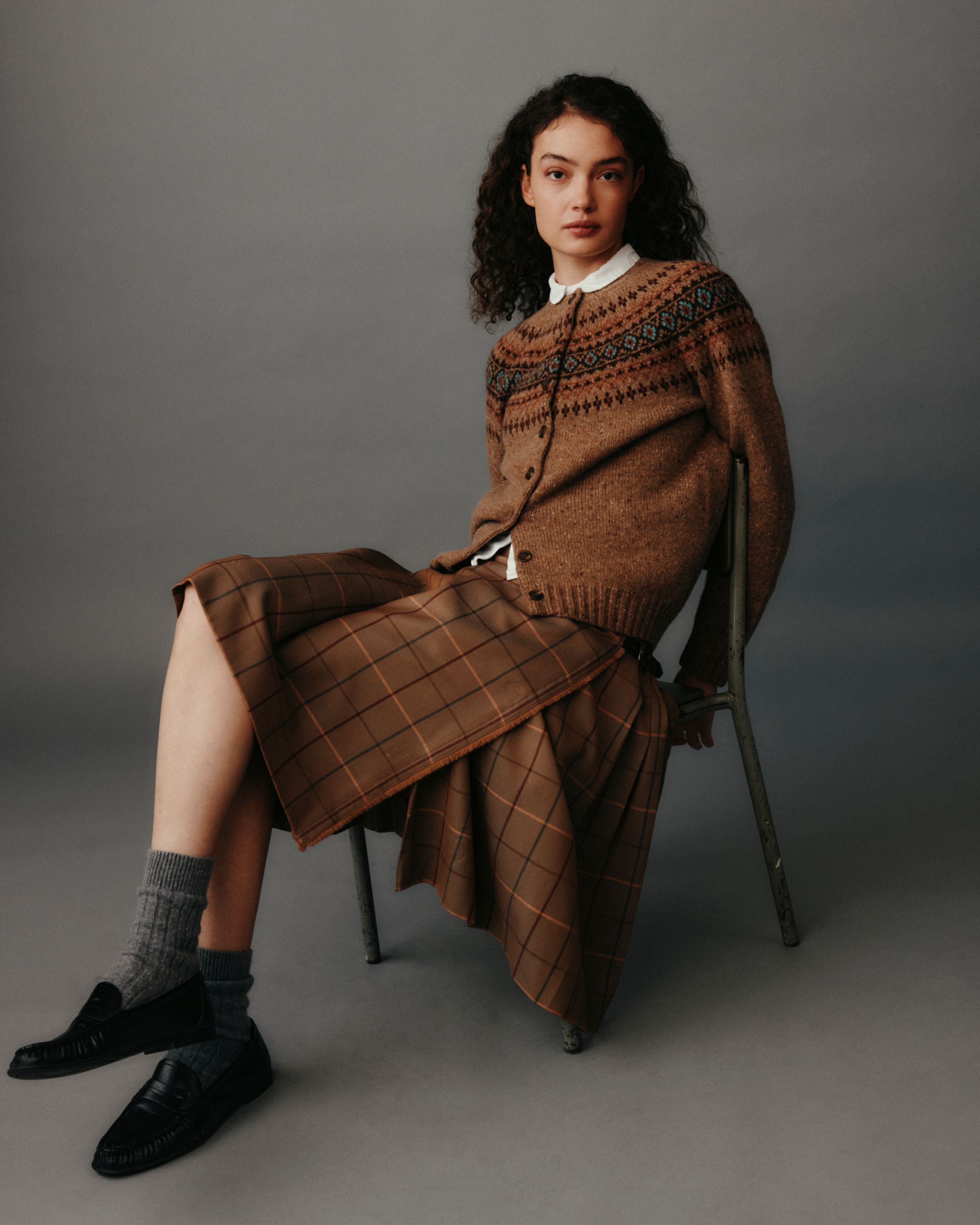 Woman wearing a brown sweater and skirt sitting on a chair against a gray background