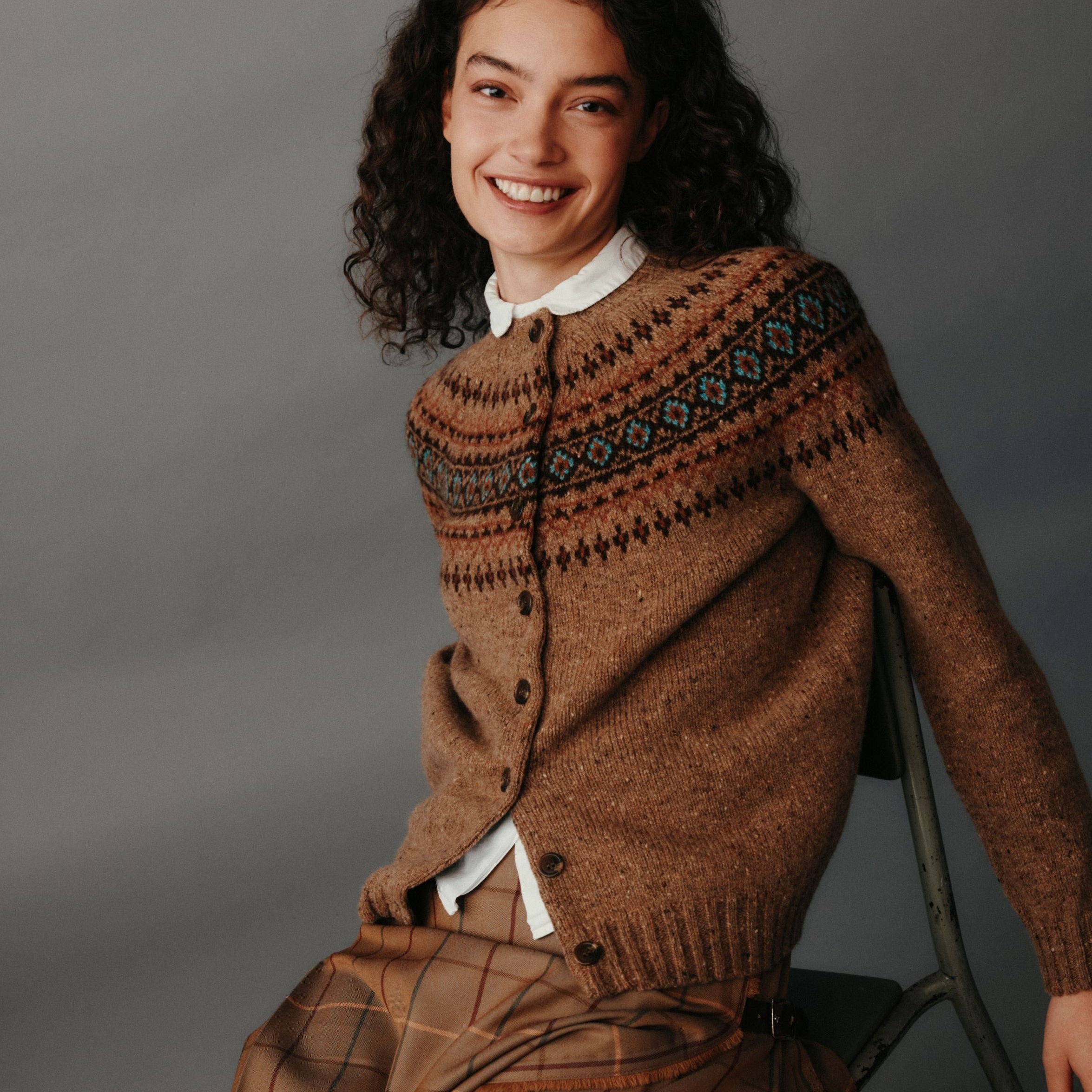 Woman wearing a brown patterned sweater and plaid skirt sitting on a chair against a gray background
