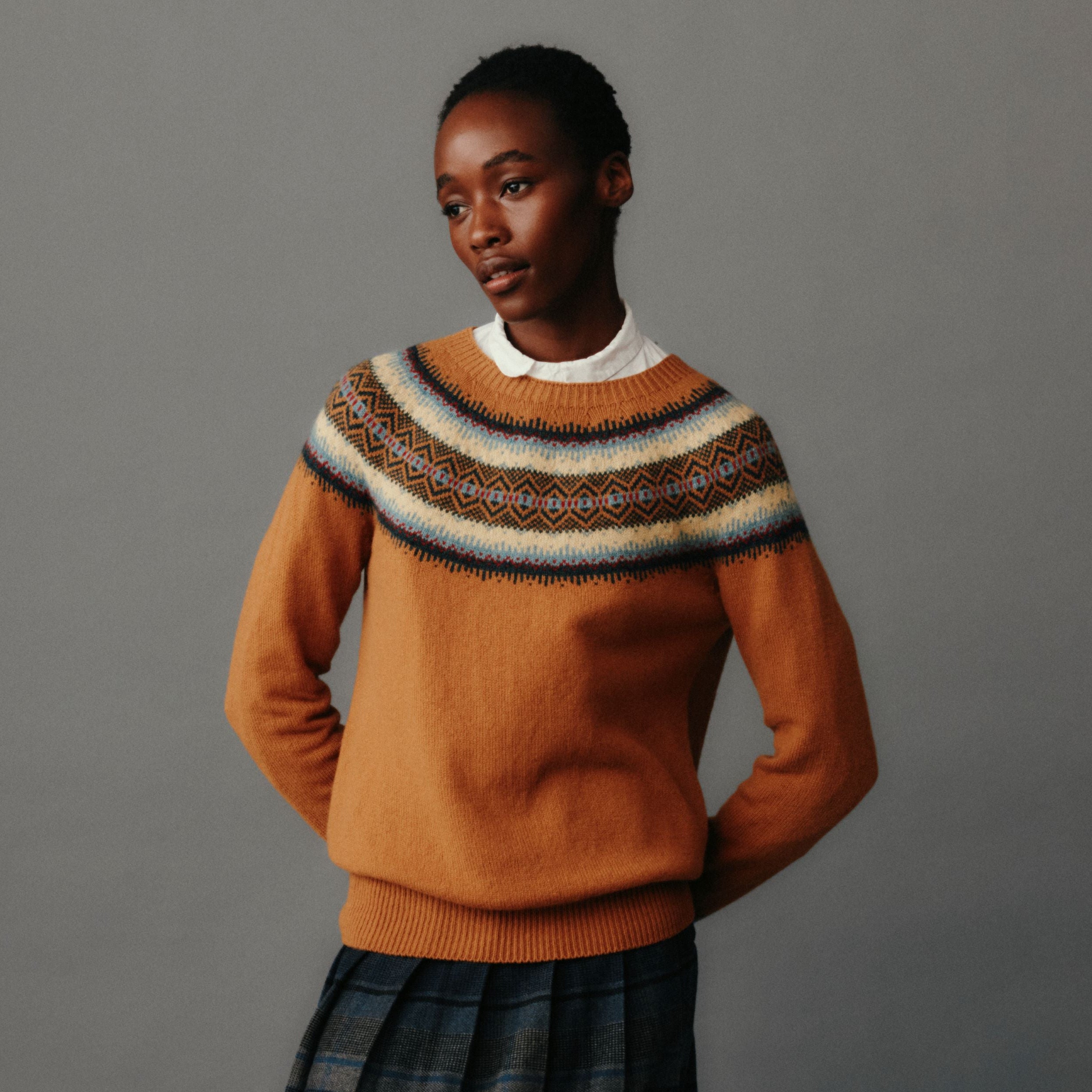 Person wearing a brown sweater with a patterned collar and plaid skirt on a gray background
