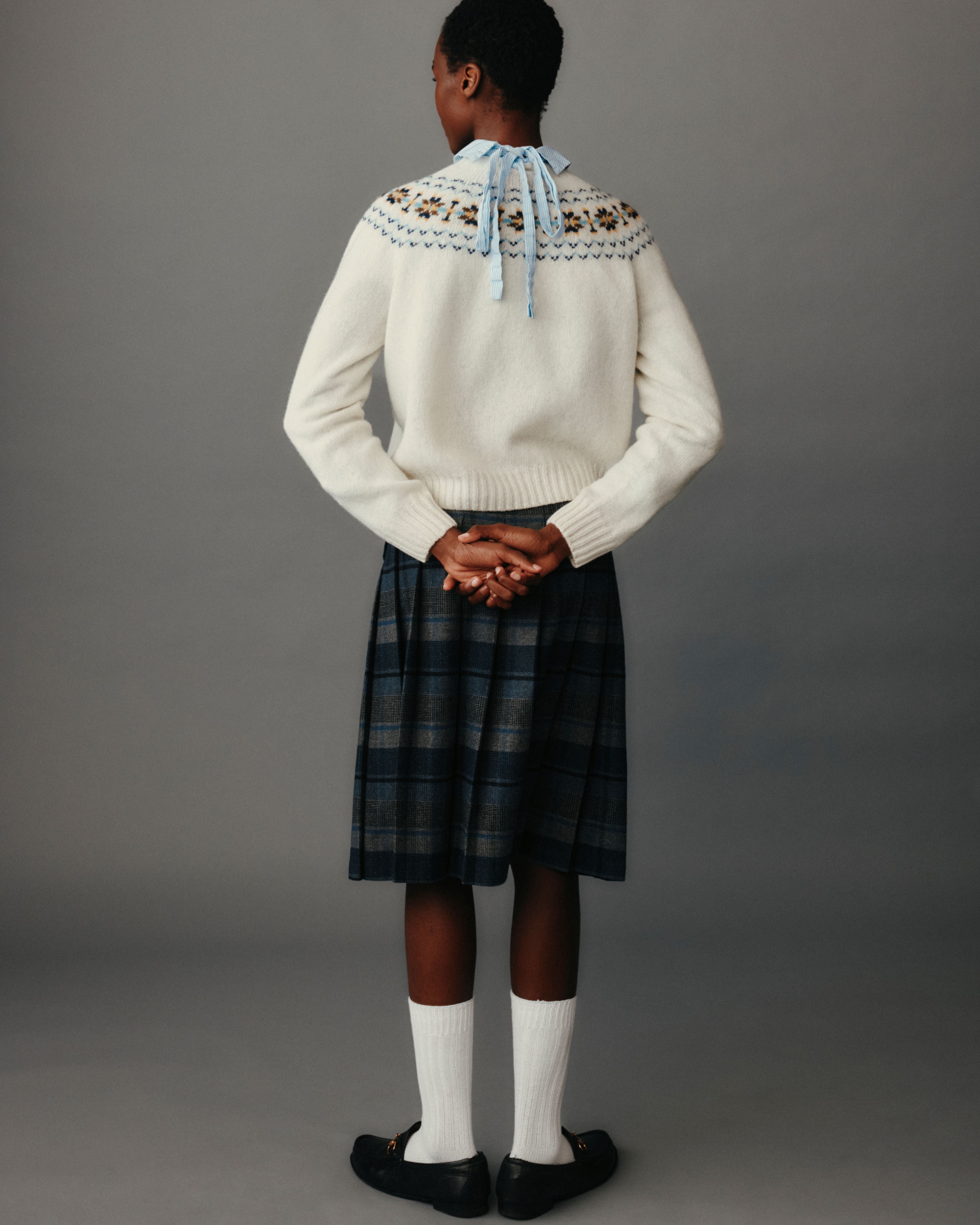 Person wearing a white sweater with patterned yoke and plaid skirt on a gray background