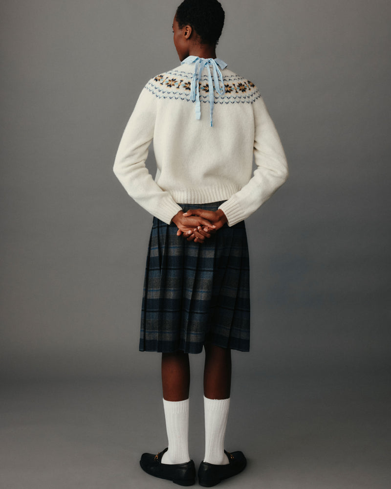 Person wearing a white sweater with patterned yoke and plaid skirt on a gray background