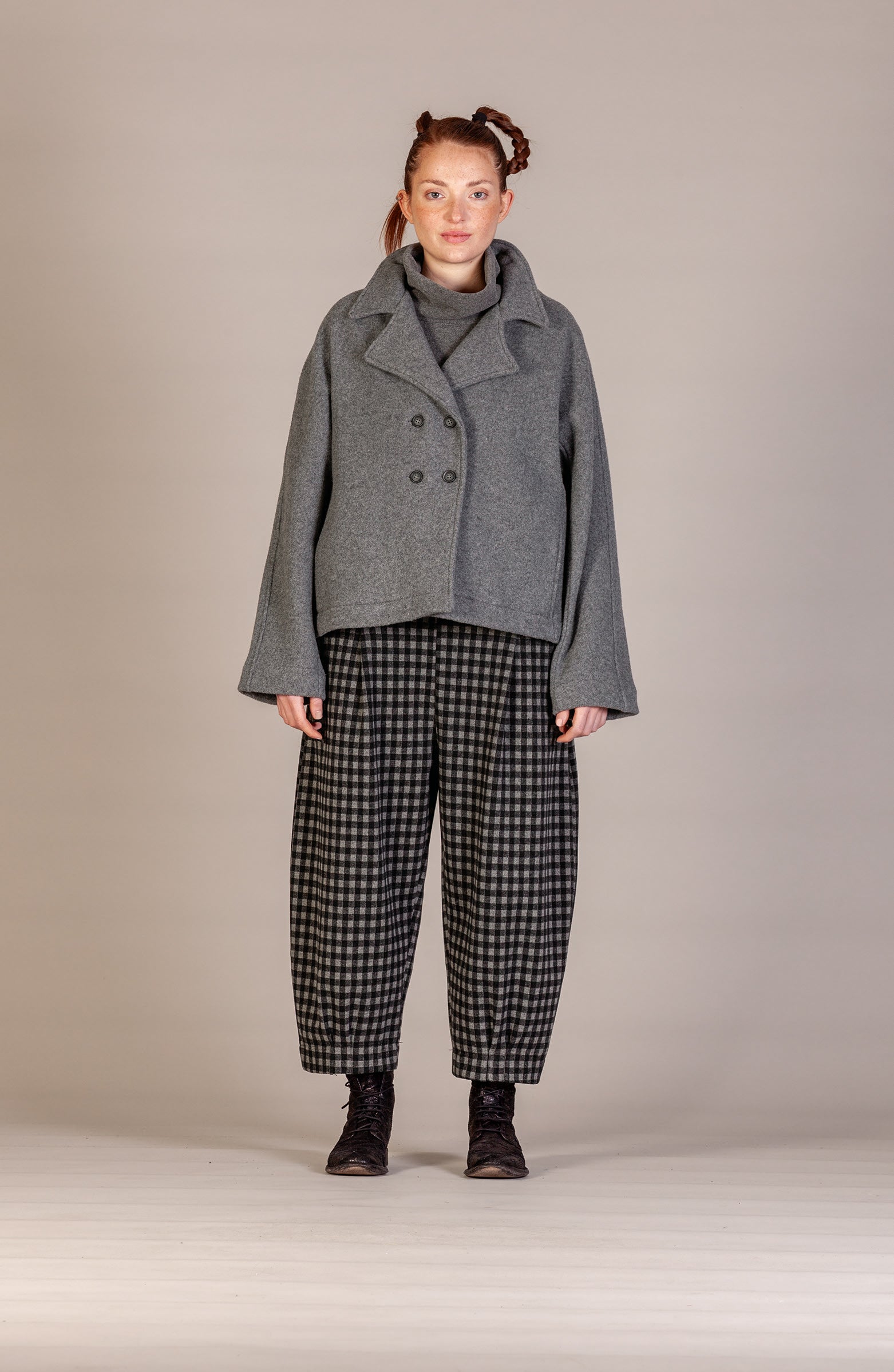 A model stands against a neutral beige studio background wearing a gray double-breasted coat over a gray turtleneck, paired with black and white checkered wide-leg trousers and dark boots.