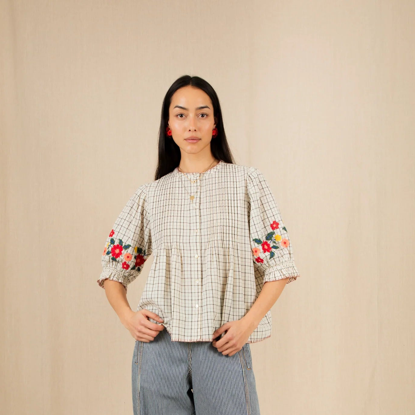 Woman wearing a checkered blouse with floral sleeves and blue pants, standing against a beige wall with flowers on the floor.