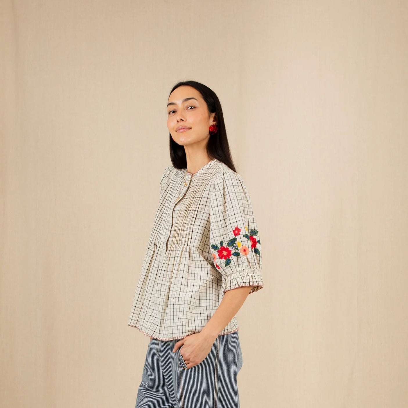Woman wearing a checkered shirt with floral embroidery and blue pants, standing against a beige wall with flowers around.