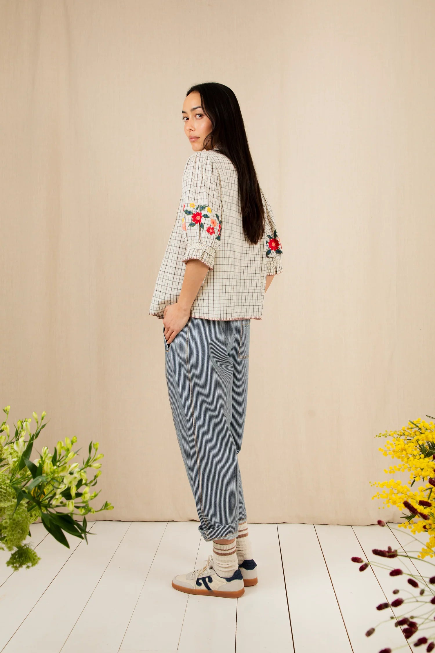 Woman wearing a checkered shirt with floral embroidery and light blue pants, standing against a beige wall with flowers on either side.