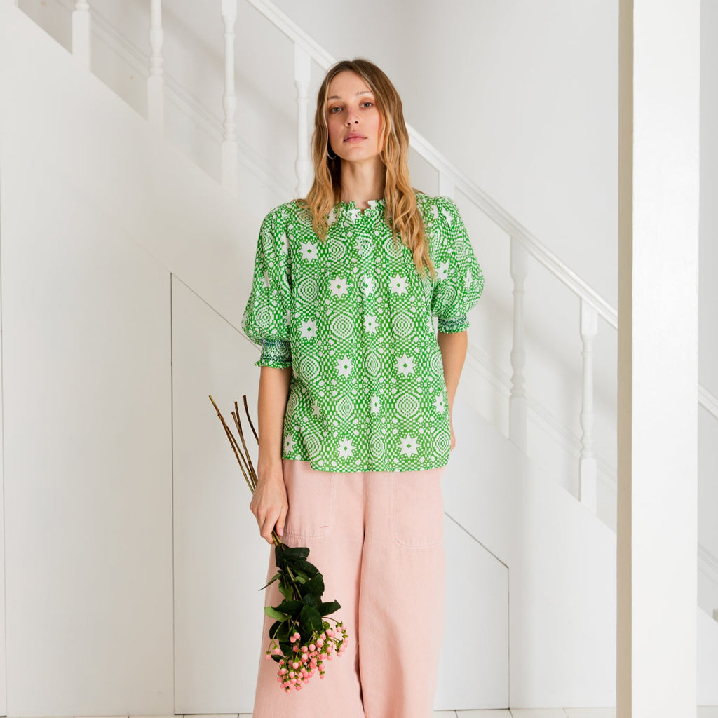 Woman wearing a green and white patterned blouse with high neck, 3/4 length sleeves with elasticated cuff. 